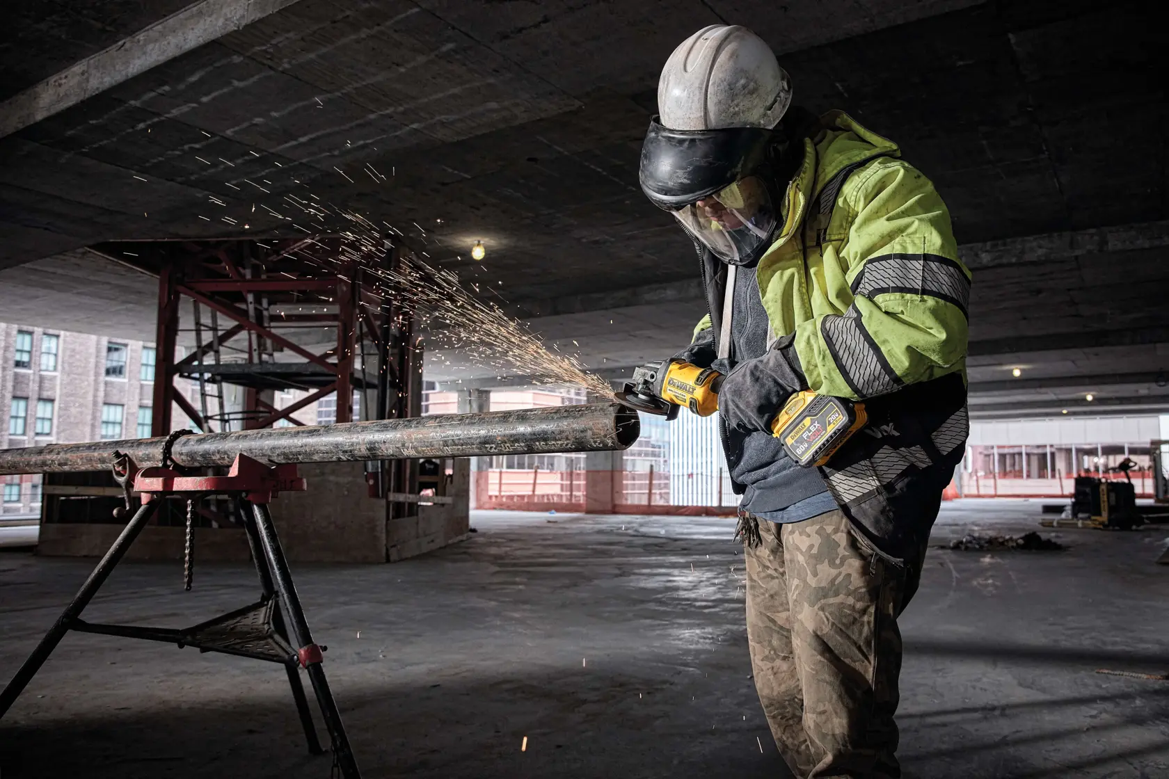 Persona con equipo de seguridad utiliza una amoladora angular inalámbrica DEWALT DCG414B para cortar un tubo de metal, produciendo chispas, en una obra de construcción.