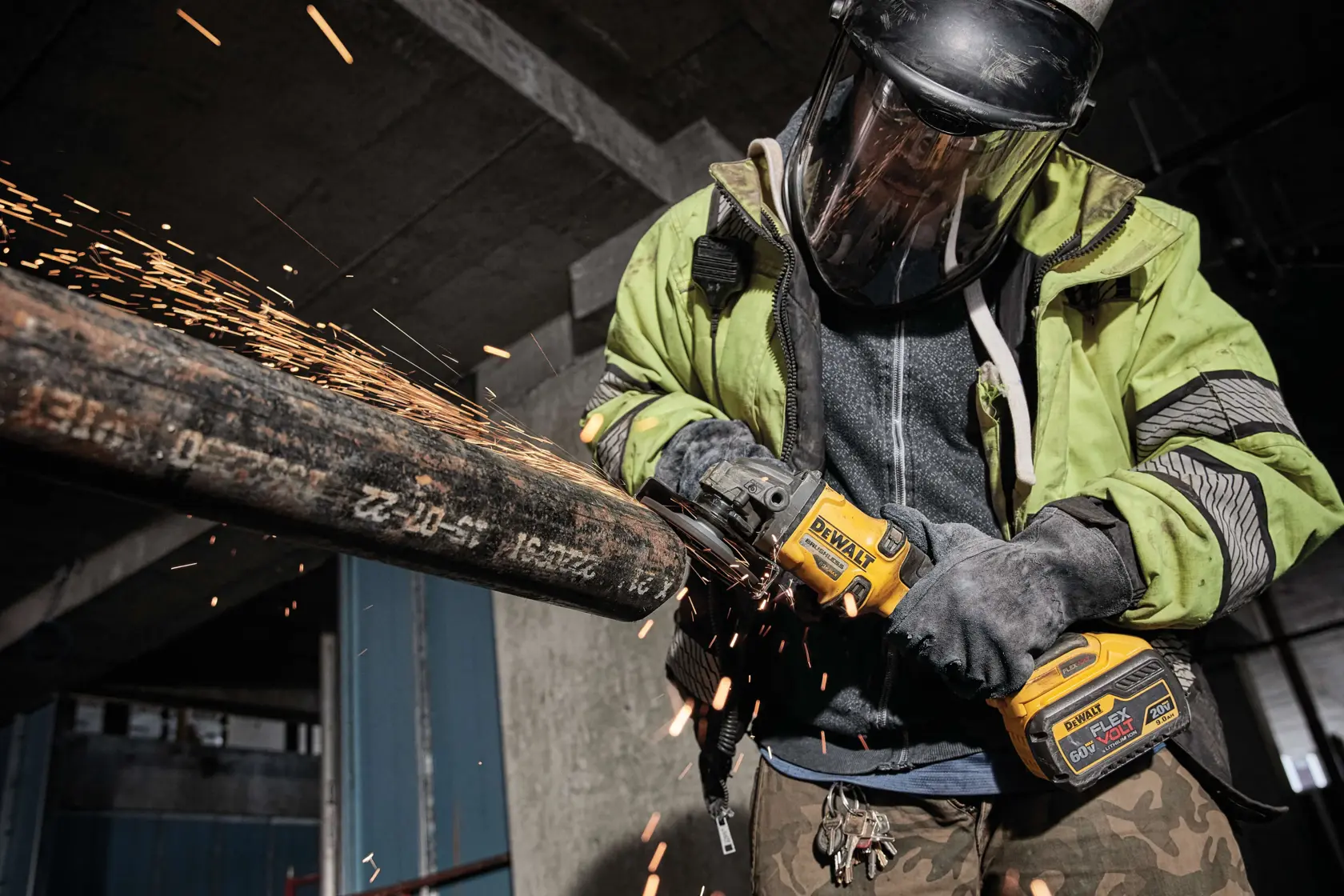 Un trabajador con equipo de protección utiliza una amoladora angular inalámbrica DEWALT (modelo DCG414B) para cortar una tubería metálica grande, generando chispas en un entorno industrial.
