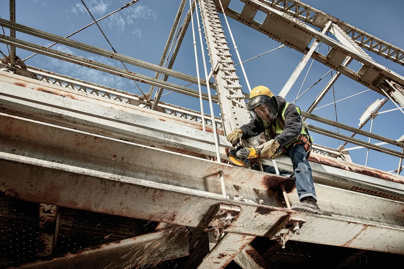Un trabajador de la construcción con equipo de seguridad utiliza una herramienta eléctrica DEWALT para cortar una viga de metal en un gran puente de acero. Se ven chispas donde la herramienta toca el metal.