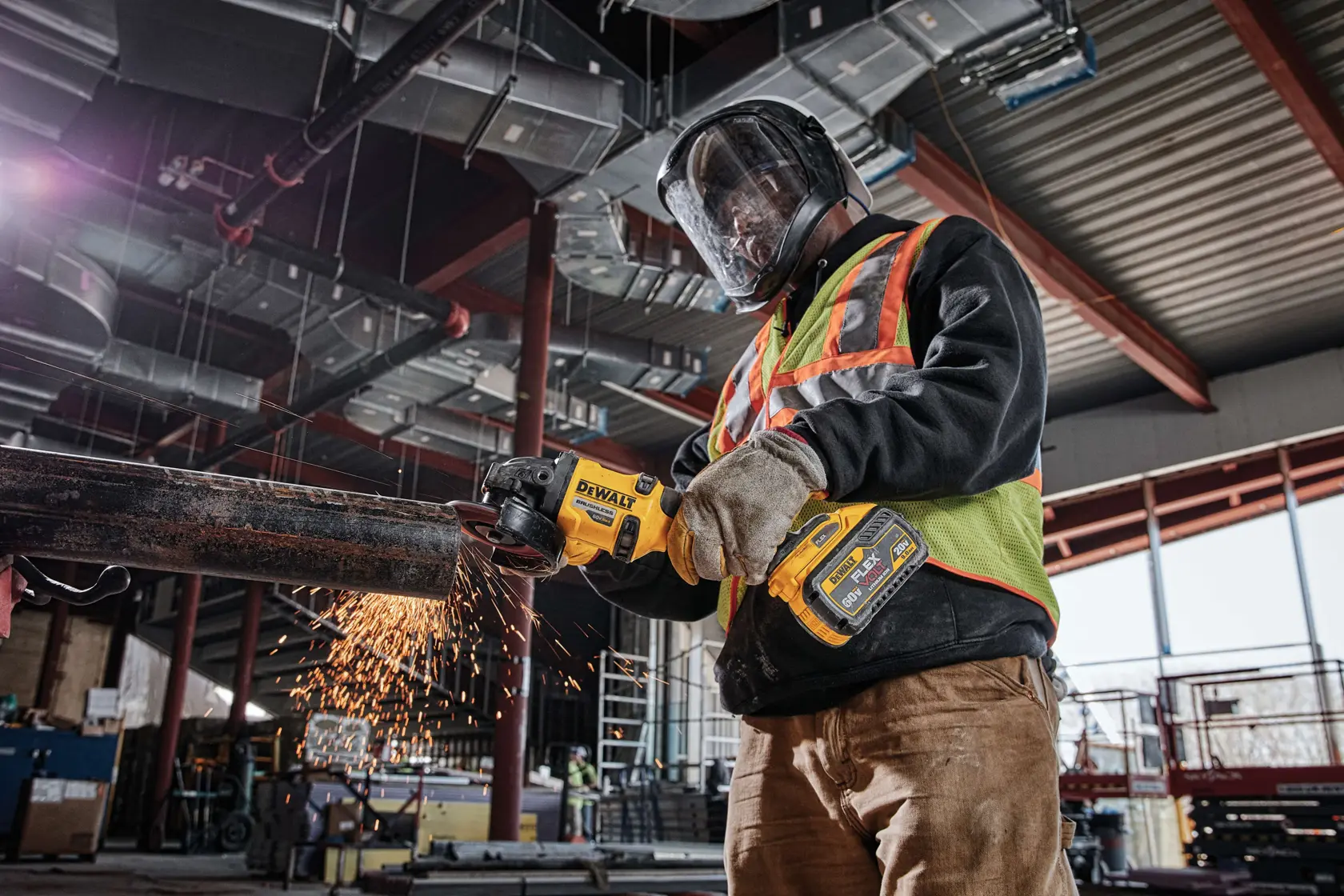 Un trabajador con equipo de protección usa una amoladora inalámbrica DEWALT DCG414B para cortar una tubería metálica, generando chispas, dentro de un edificio industrial en construcción.
