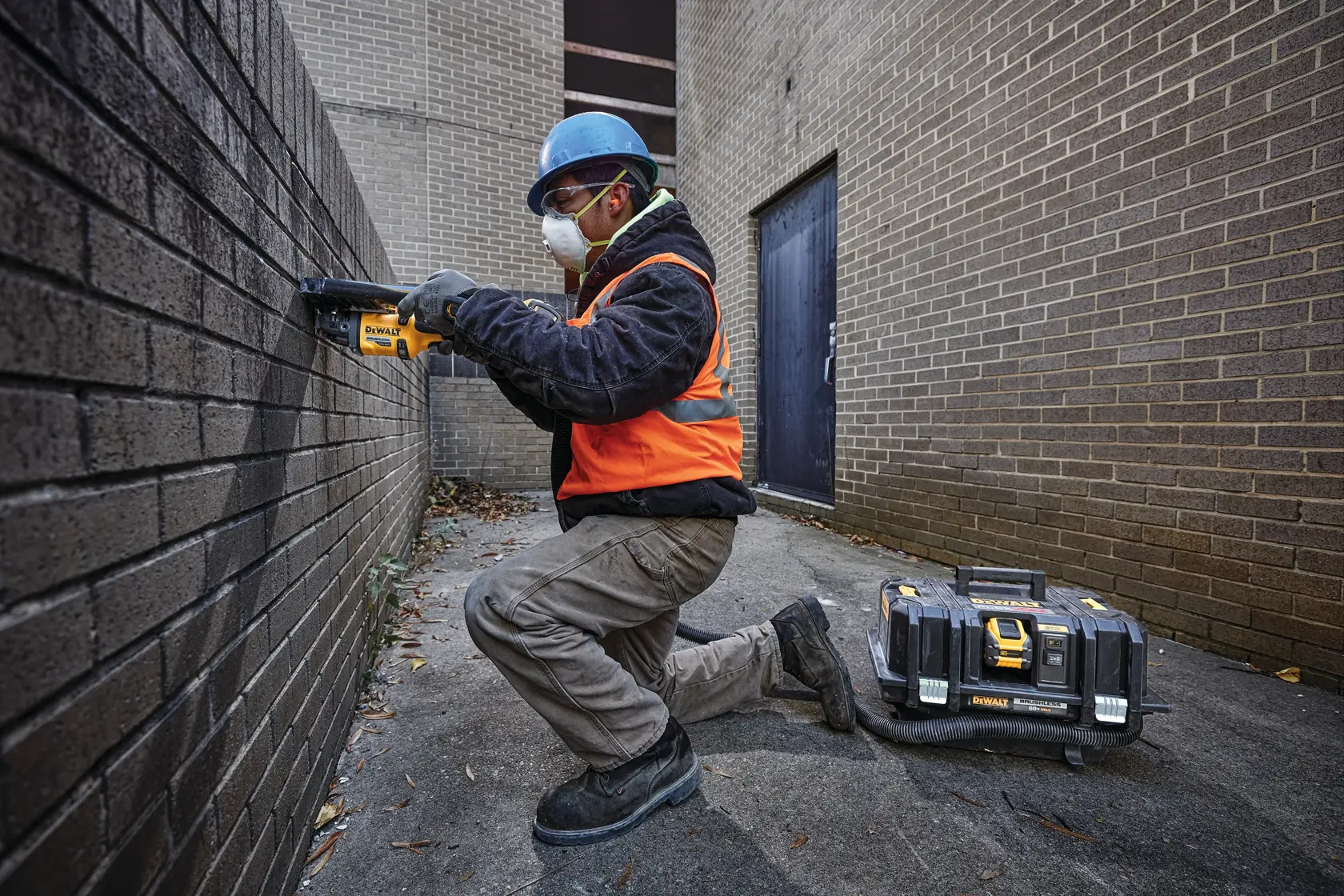 Una persona con casco azul, chaleco de seguridad naranja y ropa protectora está arrodillada al aire libre utilizando una herramienta eléctrica DEWALT en una pared de ladrillo. Un maletín negro y amarillo de DEWALT está en el suelo cerca. El rostro de la persona está difuminado por privacidad.