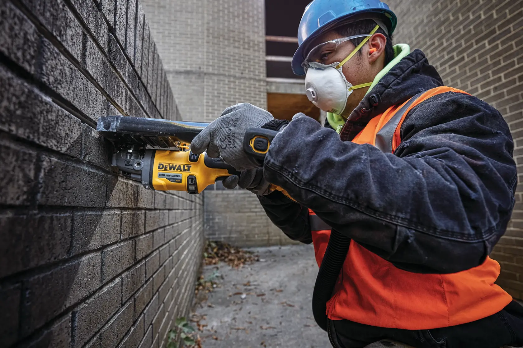 Una persona con casco de seguridad, chaleco naranja y guantes utiliza una amoladora angular inalámbrica DEWALT para cortar una pared de ladrillos en una obra.