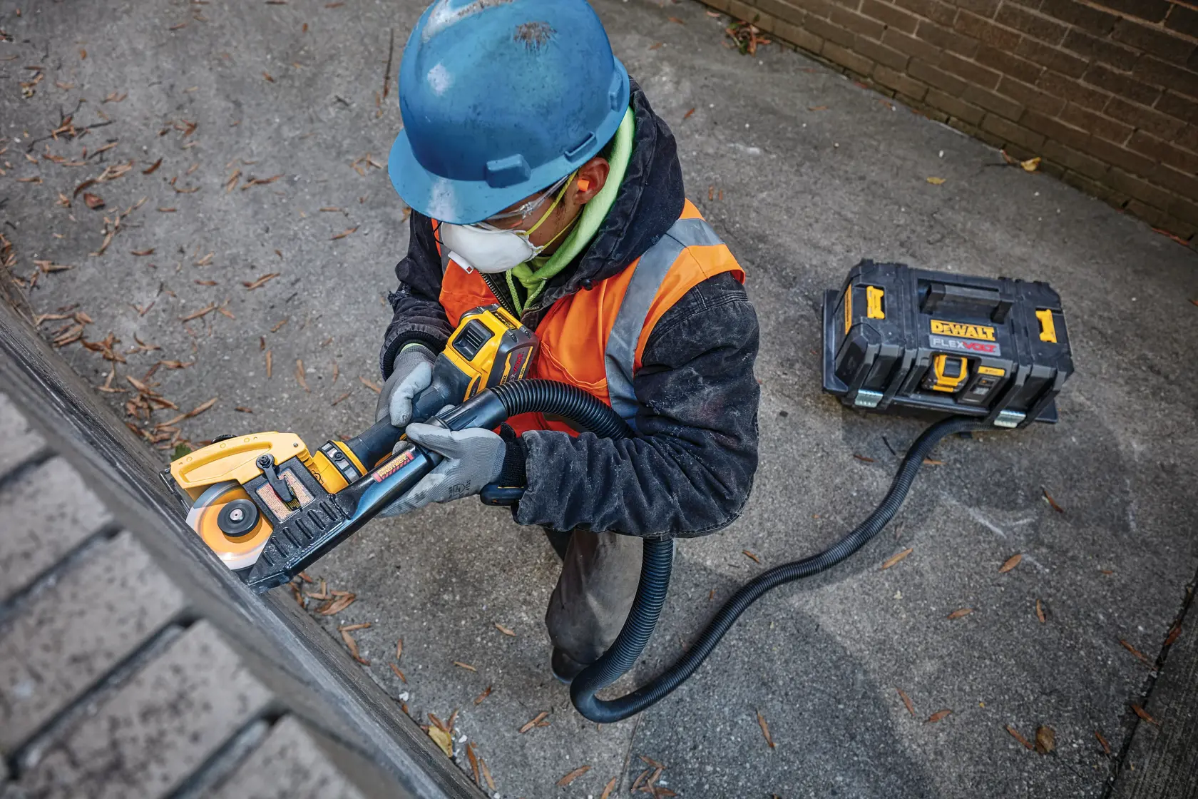 Un trabajador con casco azul y chaleco de seguridad naranja utiliza una herramienta eléctrica DEWALT con una unidad de extracción de polvo conectada por una manguera, trabajando sobre una superficie de ladrillo al aire libre.