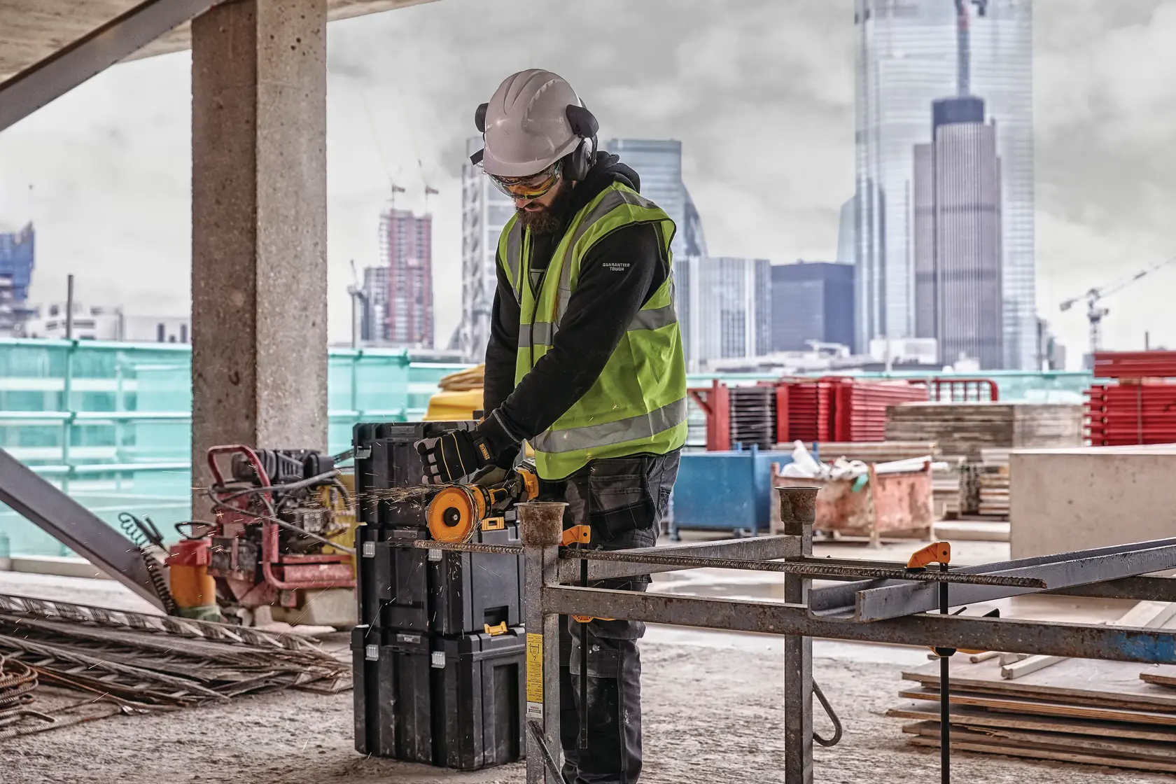 Un trabajador de construcción con chaleco reflectante, casco y protección auditiva utiliza una herramienta eléctrica portátil Dewalt en un sitio de construcción, con rascacielos de la ciudad al fondo. Hay varios materiales y equipos de construcción alrededor.