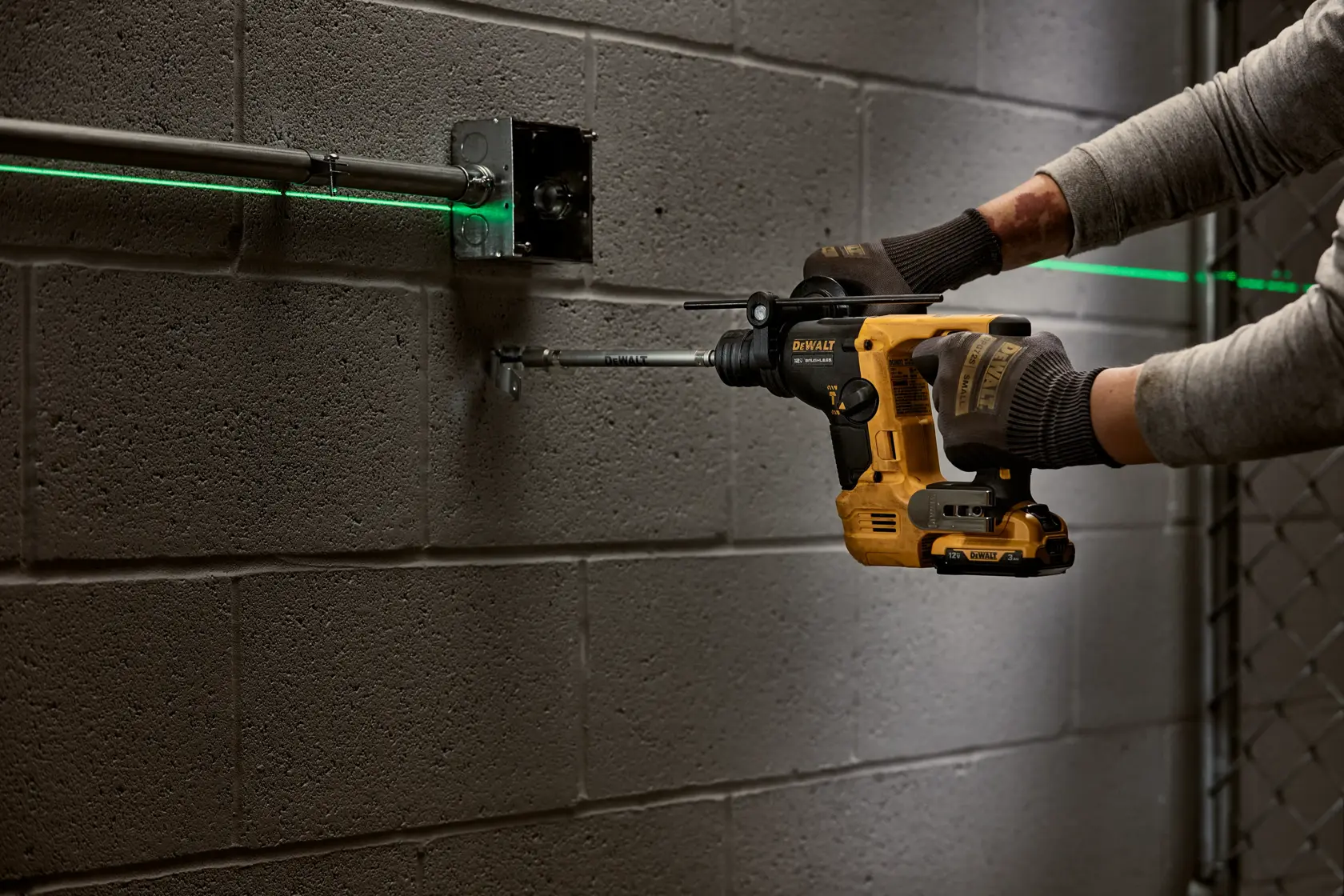 Una persona con guantes utiliza un martillo rotativo inalámbrico amarillo DEWALT para perforar una pared de bloques de hormigón, alineando un conducto eléctrico con la ayuda de un nivel láser verde.