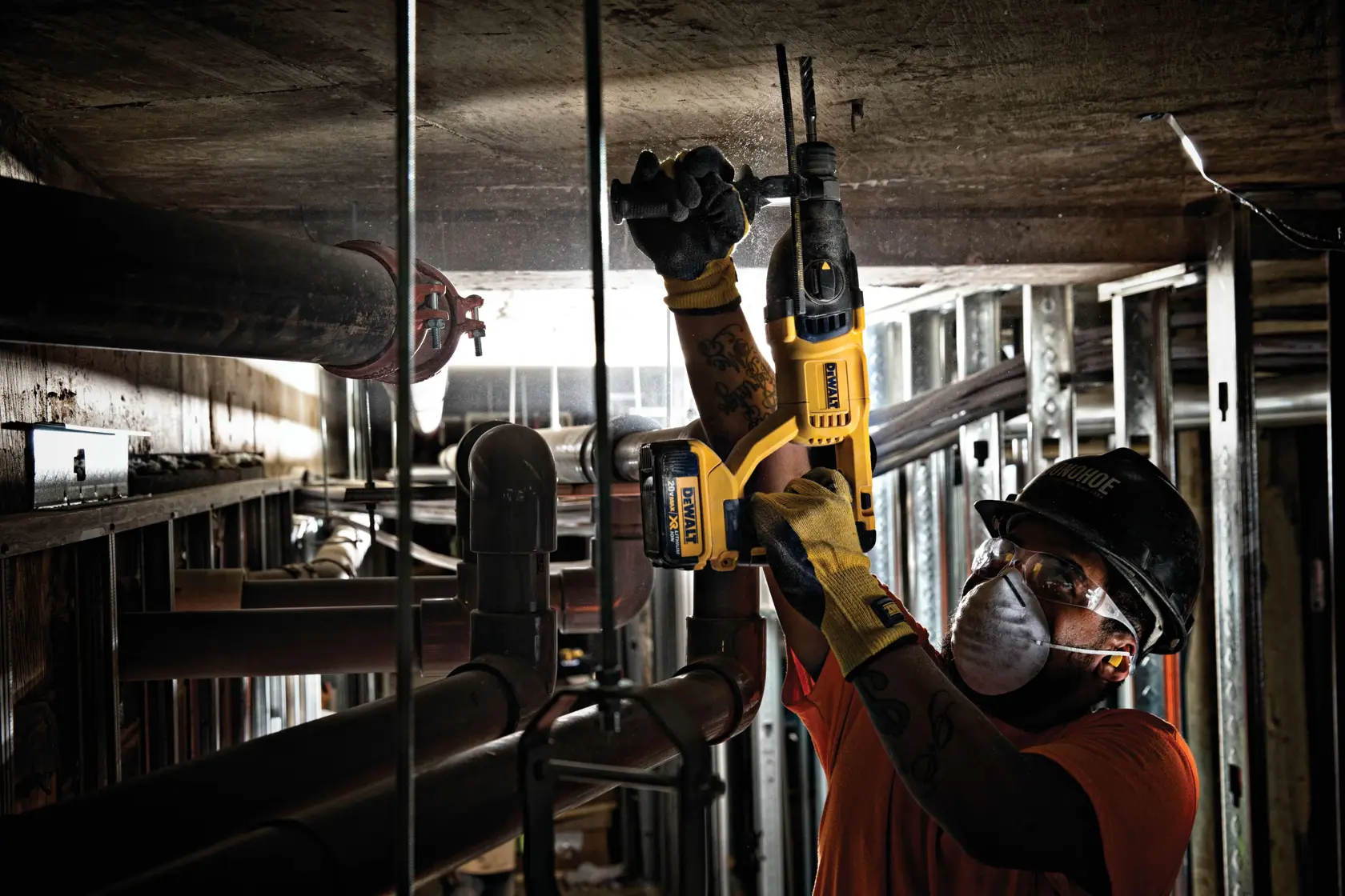 Un trabajador de la construcción con guantes y casco utiliza un martillo rotativo inalámbrico amarillo DEWALT para trabajar en un techo de concreto en un entorno industrial con tuberías y estructuras metálicas expuestas.