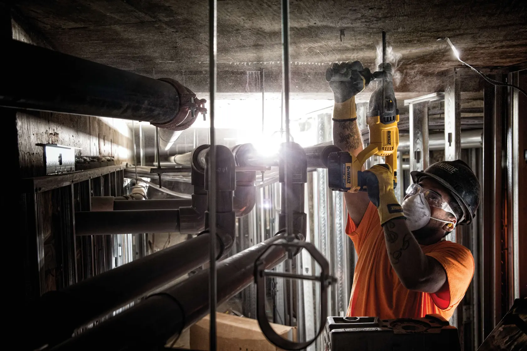 Un trabajador con equipo de protección utiliza un taladro rotativo inalámbrico amarillo DEWALT (modelo DCH133B) para perforar un techo de concreto en un entorno industrial con tuberías metálicas y materiales de construcción visibles.