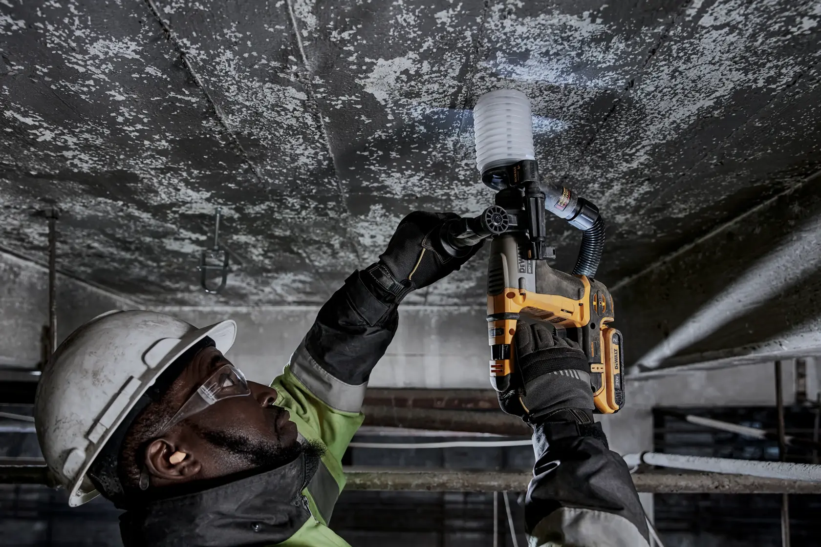 Una persona con equipo de seguridad y guantes está usando un martillo rotatorio inalámbrico DEWALT (modelo DCH172B) para perforar un techo de concreto. La herramienta está equipada con un accesorio de extracción de polvo.