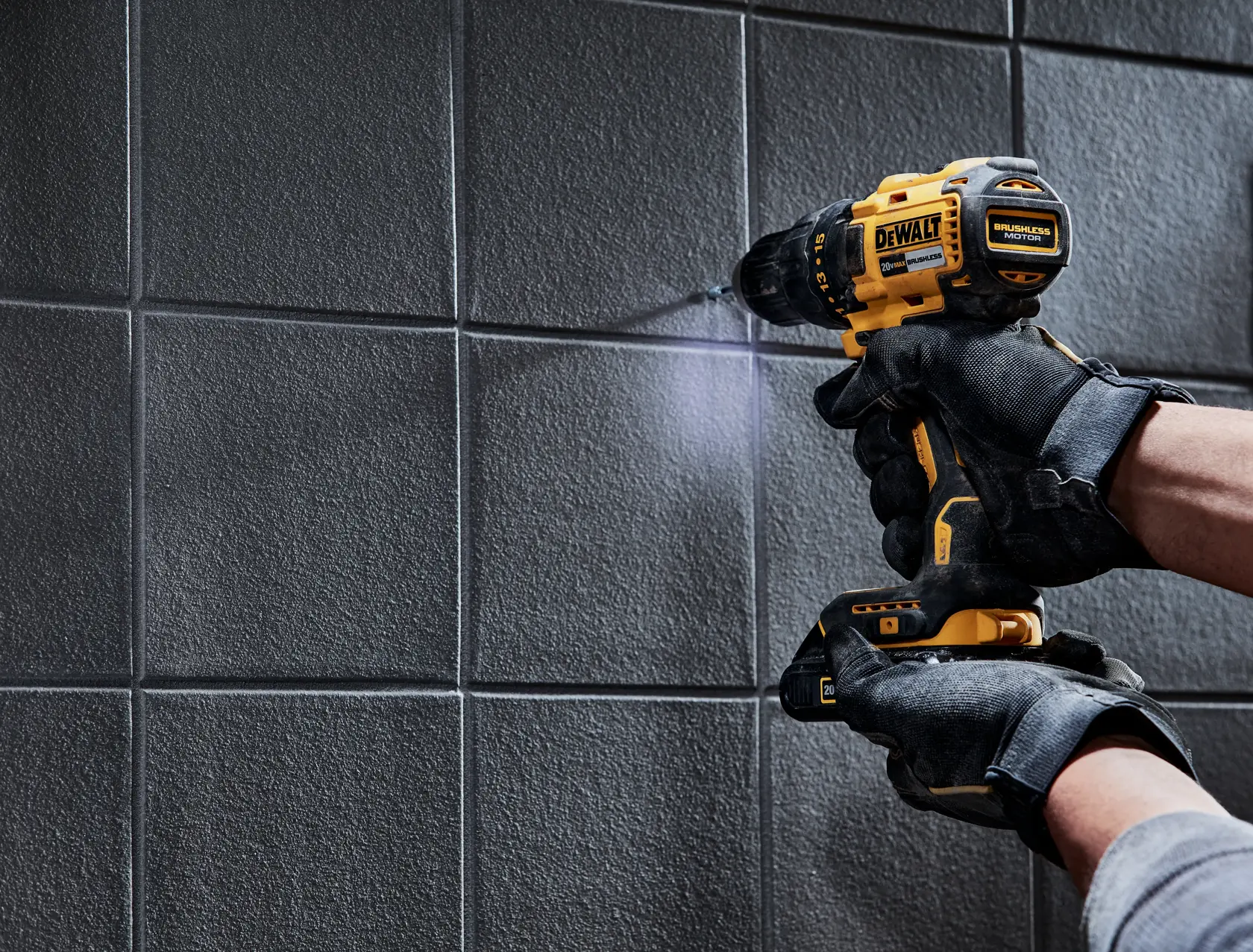 Una persona con guantes negros está usando un taladro inalámbrico amarillo DEWALT para perforar una pared de baldosas texturizadas.