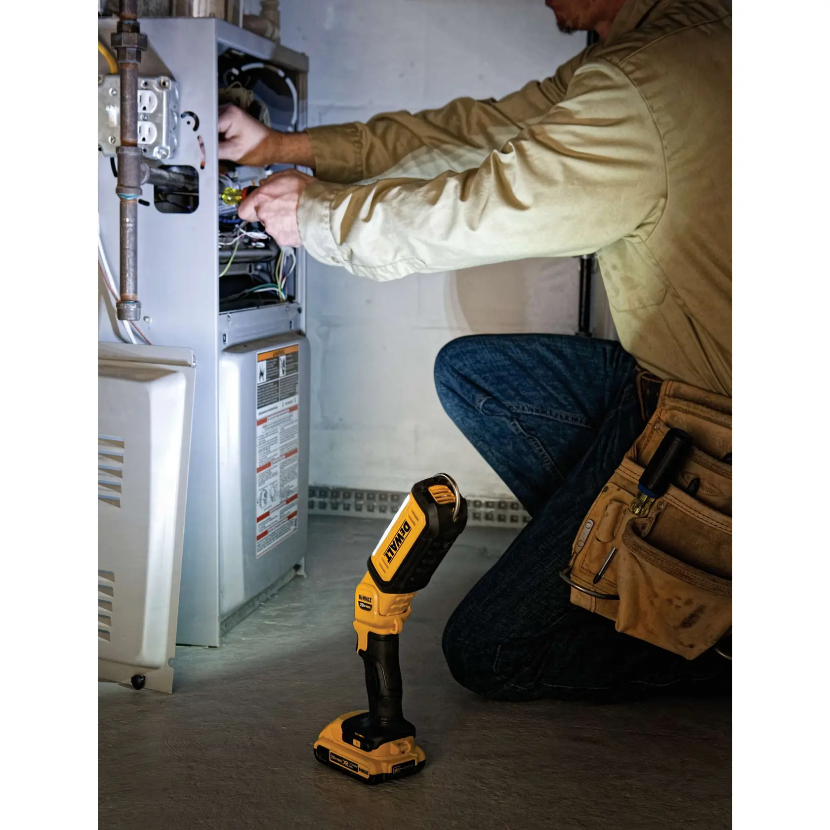 Una persona está arrodillada y trabajando en una unidad eléctrica abierta, utilizando herramientas. En el suelo hay una lámpara de trabajo LED portátil DEWALT DCL050 que ilumina el área de trabajo.