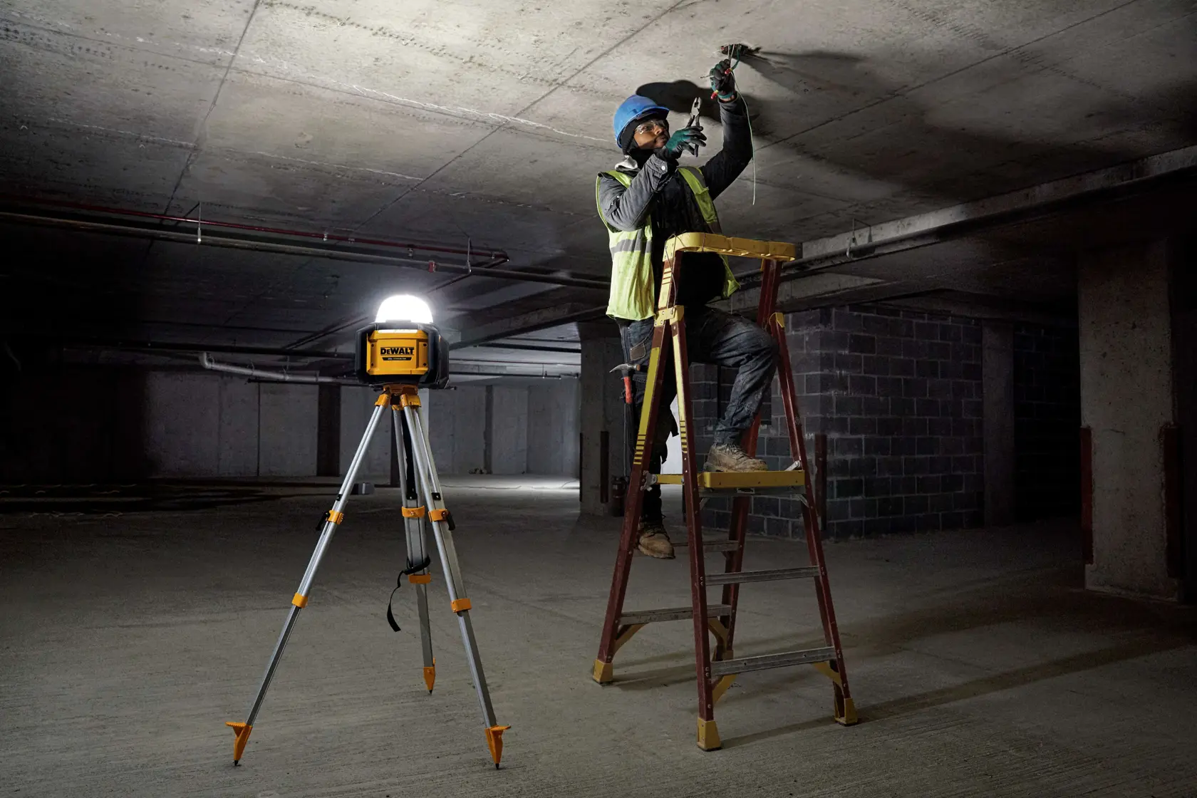 Una luz de trabajo portátil DEWALT montada en un trípode ilumina un sitio de construcción oscuro. Cerca, un trabajador con equipo de seguridad está de pie en una escalera y trabaja en el techo.