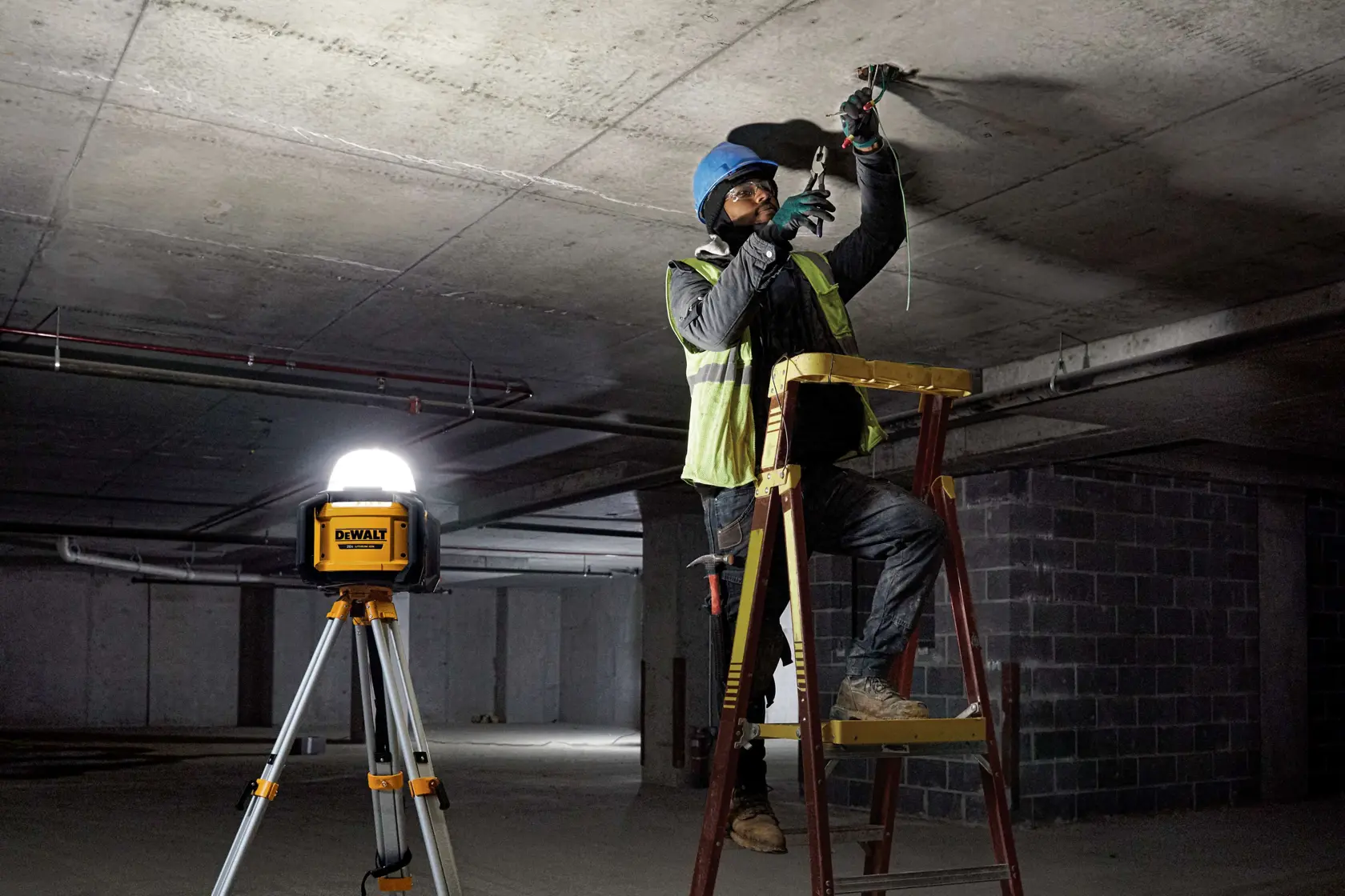 Una lámpara de trabajo DEWALT DCL074 sobre un trípode brinda iluminación a un trabajador de la construcción en una escalera, trabajando en el techo de un edificio sin terminar.
