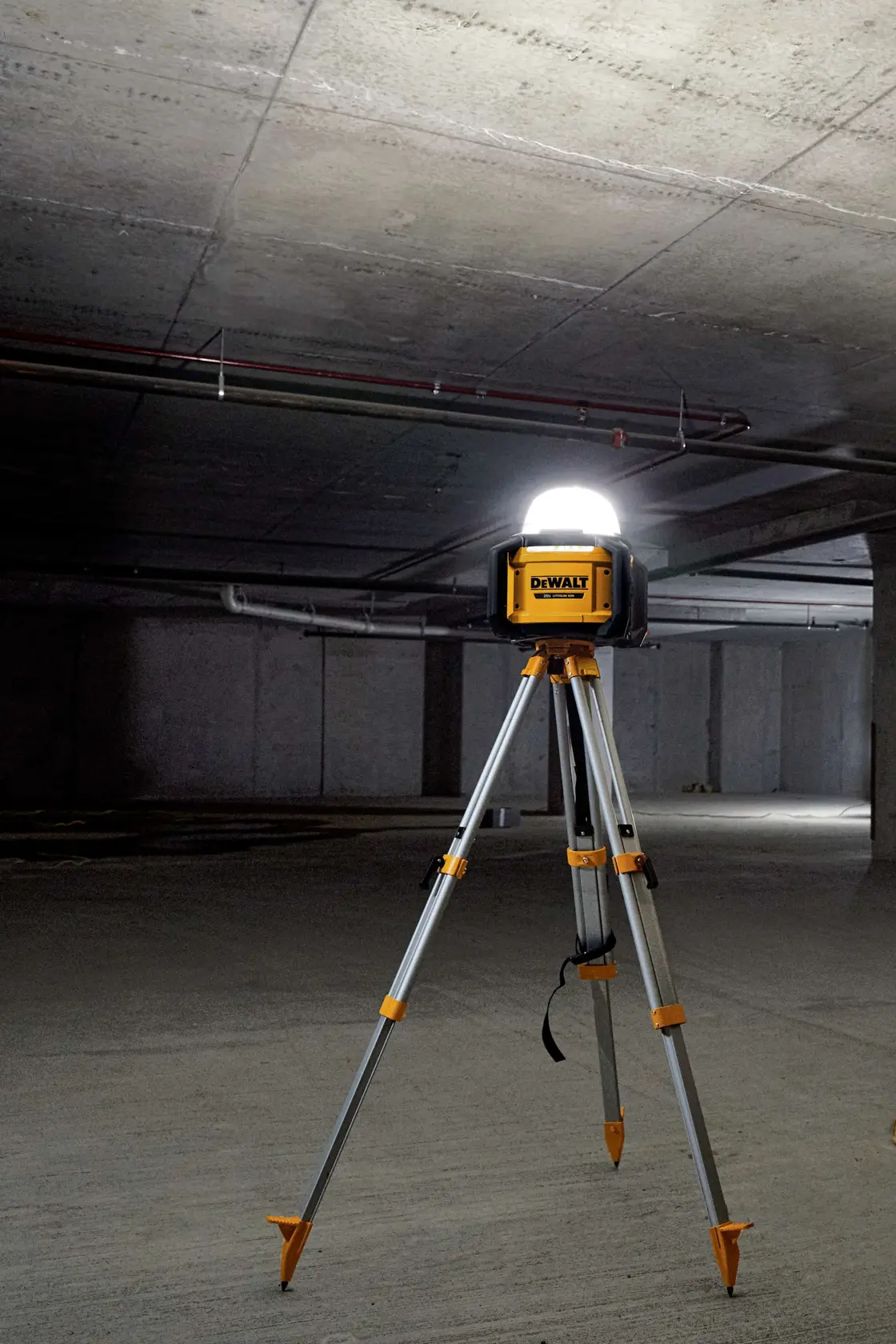 Una luz de trabajo portátil DEWALT sobre un trípode, iluminando una habitación grande vacía de concreto o un sótano.