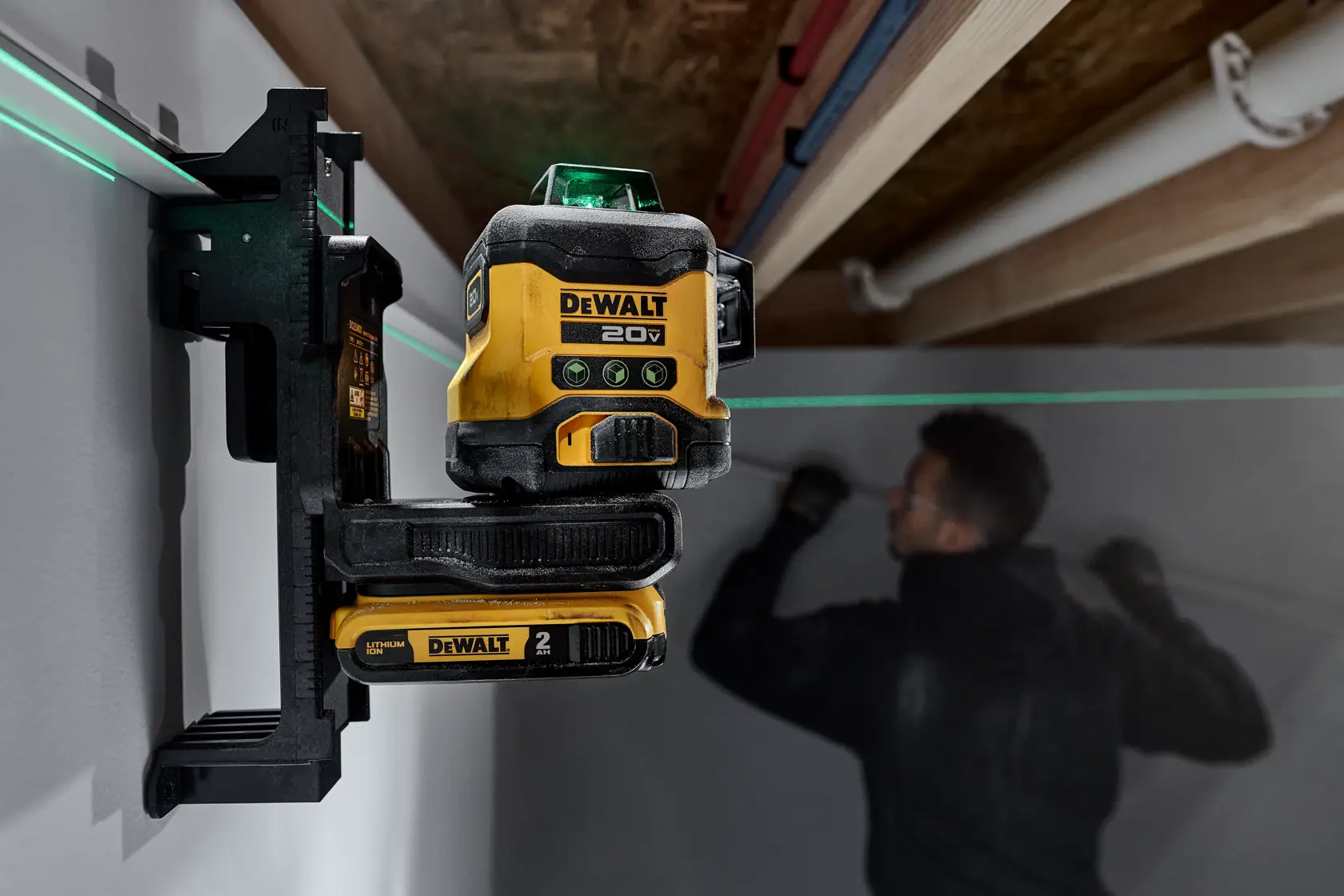 Un nivel láser DEWALT 20V está montado en una pared, proyectando una línea láser horizontal verde. Una persona está trabajando en el fondo sobre la pared, con el rostro borroso.