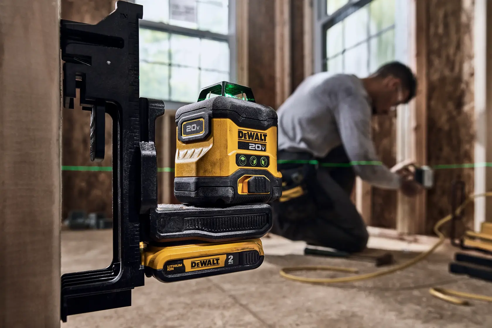 Una herramienta niveladora láser DEWALT 20V montada en una abrazadera, proyectando líneas láser verdes en un sitio de construcción. Una persona trabaja en el fondo sobre una estructura de madera.