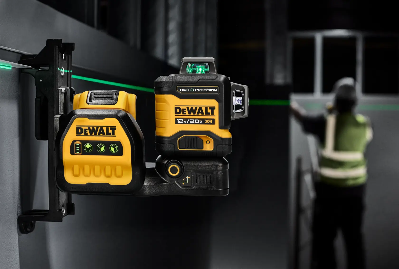 Un nivel láser verde de alta precisión DEWALT 12V/20V XR está montado en un soporte de pared. Emite una línea láser verde visible a través del área de trabajo. El dispositivo es amarillo y negro con la marca DEWALT. Al fondo, una persona con chaleco reflectante trabaja cerca de la línea láser.