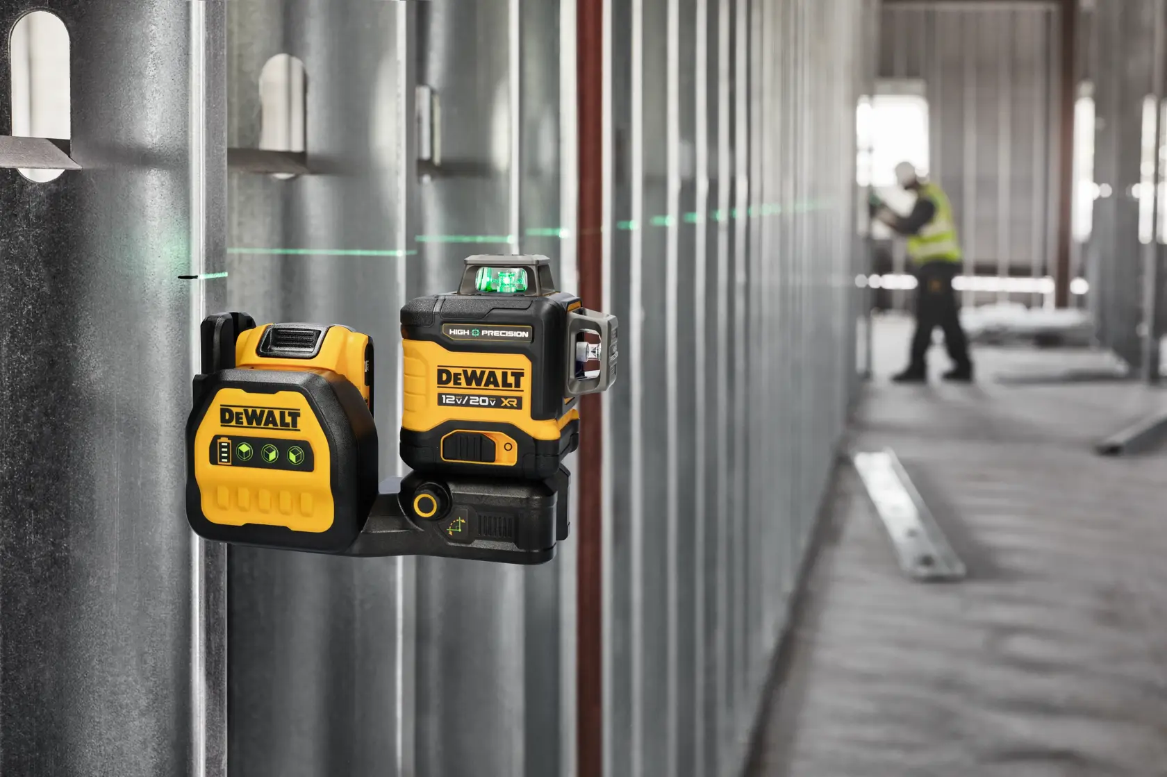 Un nivel láser DEWALT está montado en una pared de metal de construcción, proyectando una línea láser horizontal verde a través de la habitación. En el fondo, una persona con chaleco de seguridad trabaja en un espacio industrial sin terminar.