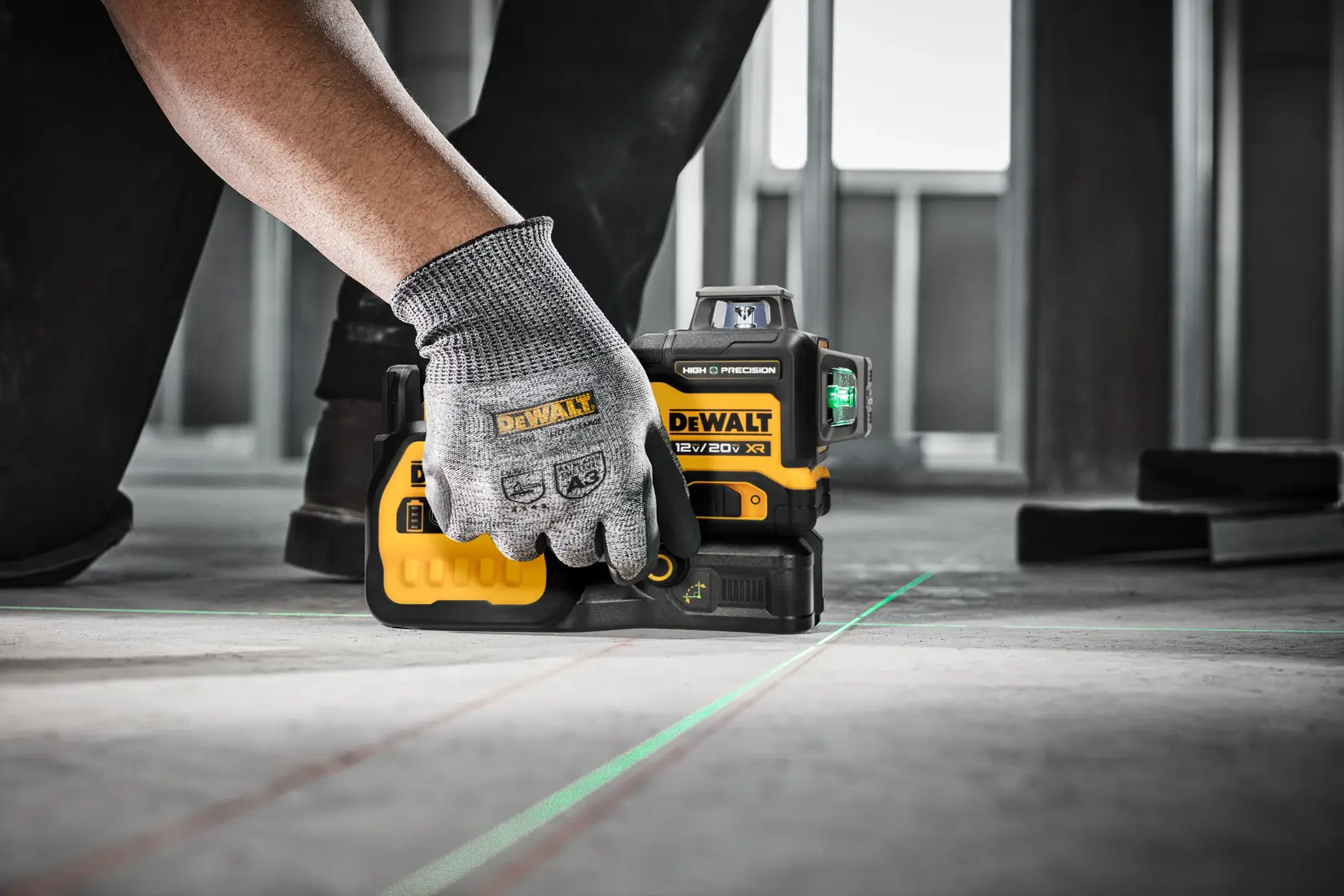 Una persona con un guante de trabajo gris está colocando un nivel láser DEWALT 12V/20V XR en un suelo de concreto, proyectando líneas láser verdes para ayudar en la alineación. La herramienta es ideal para tareas de precisión en la construcción.
