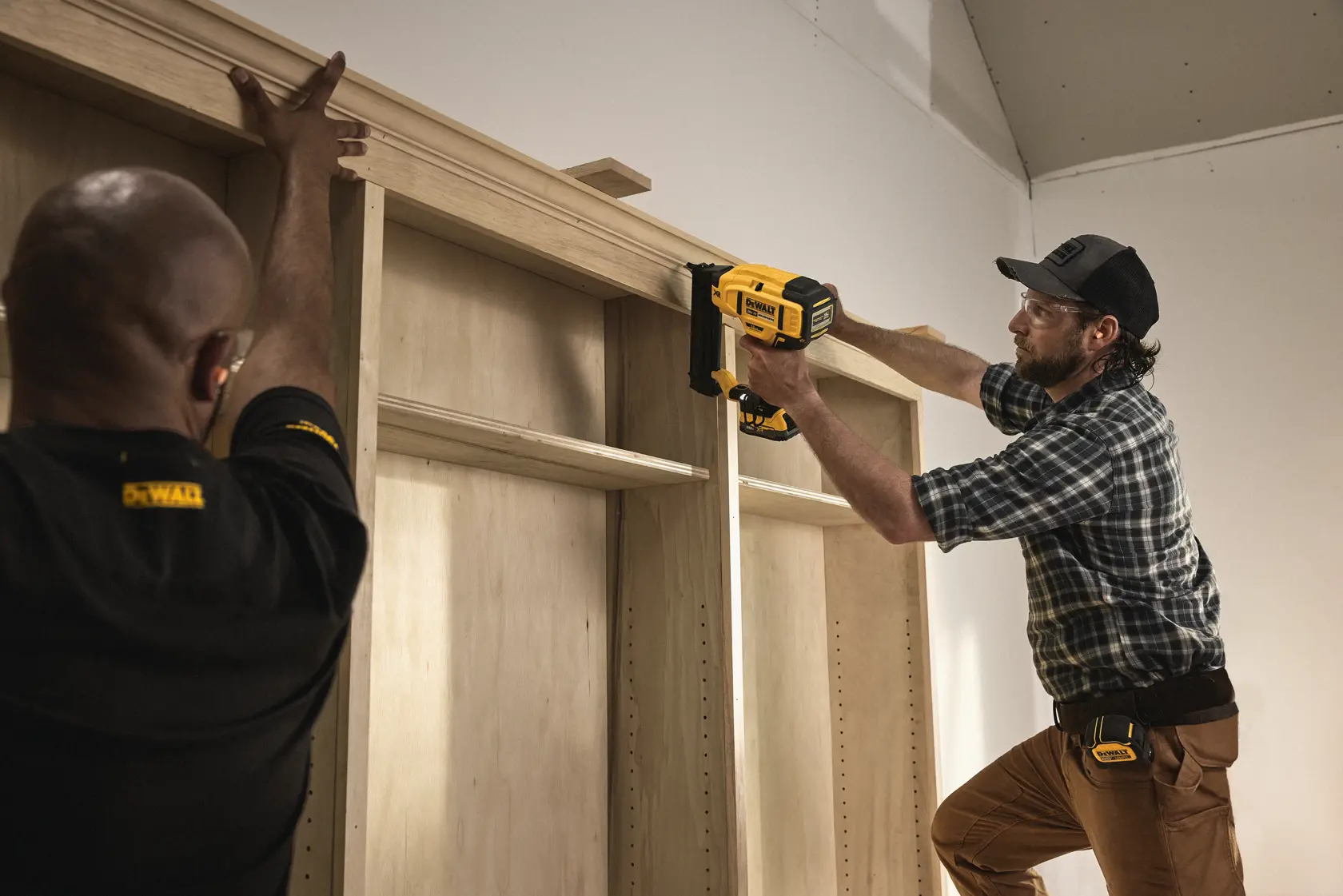 Dos personas están instalando un adorno de madera en un gabinete usando una clavadora inalámbrica DEWALT. Una persona sostiene la madera mientras la otra utiliza la clavadora.