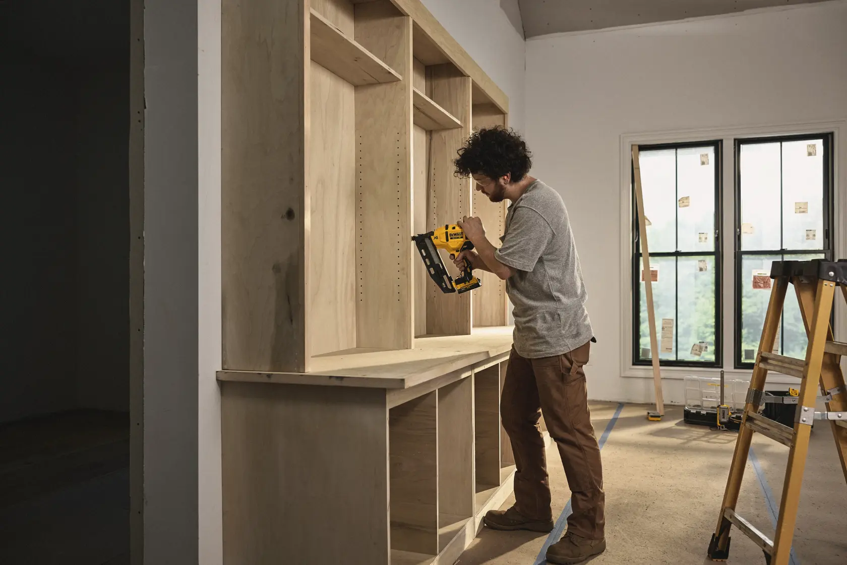 Una persona está utilizando una clavadora inalámbrica DEWALT para ensamblar estanterías de madera en una habitación en construcción. La persona está de pie cerca de una escalera y una ventana, vistiendo ropa de trabajo.