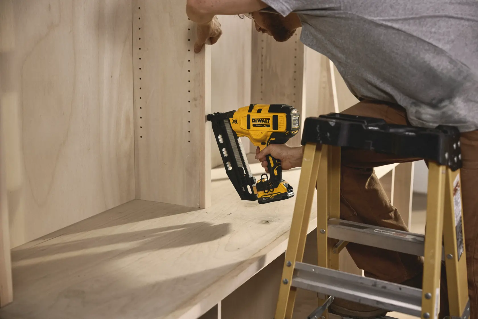 Una persona utiliza una pistola de clavos inalámbrica DEWALT (modelo DCN660B) para fijar un panel de madera dentro de una unidad de estantes. La persona está de pie sobre una escalera y trabaja cuidadosamente con la herramienta.