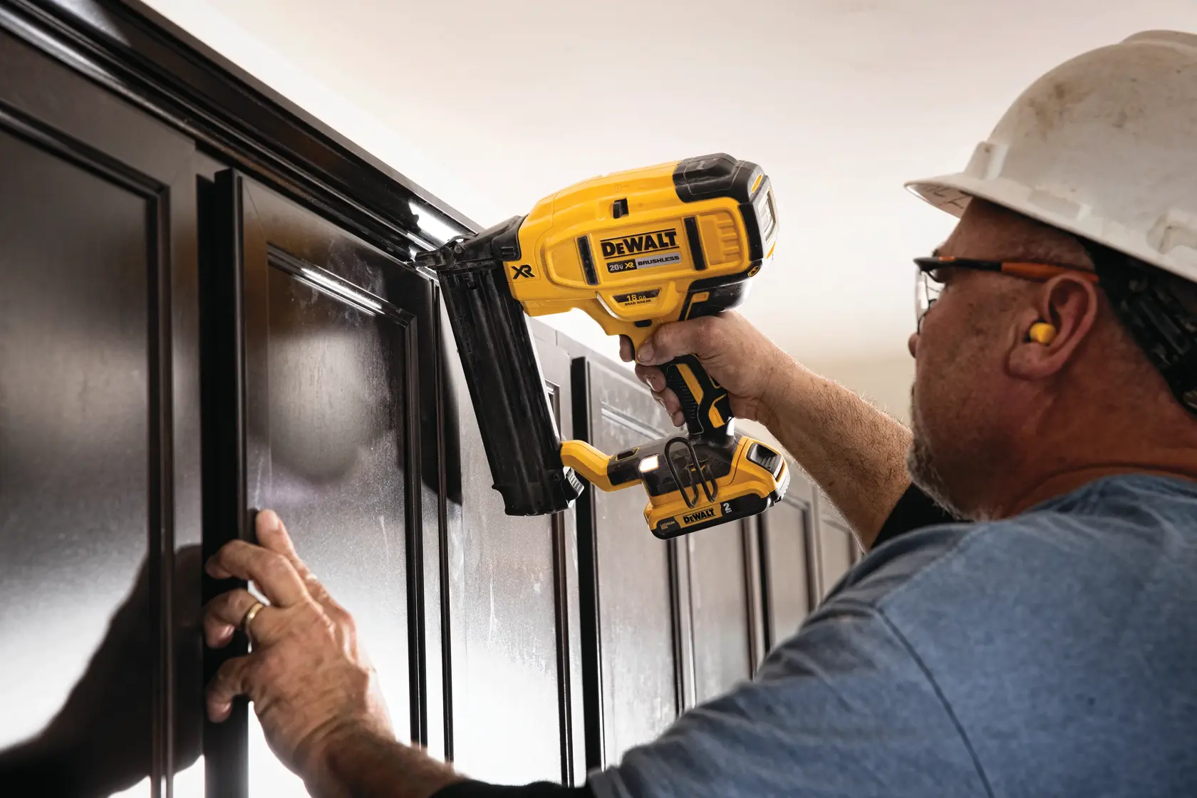 Una persona con equipo de seguridad utiliza una clavadora inalámbrica DEWALT para instalar molduras en gabinetes de madera oscura.