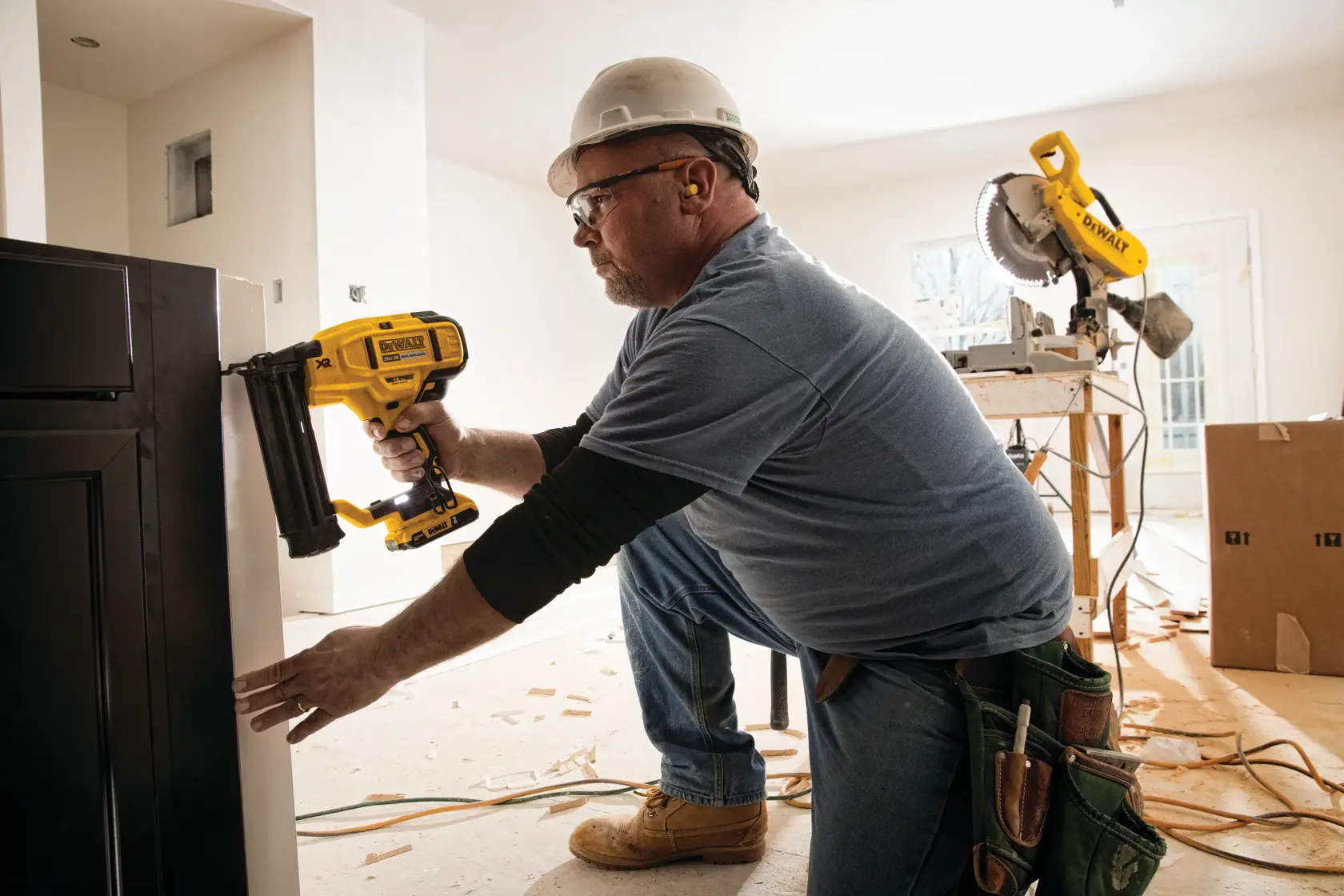 Una persona con casco y protección auditiva utiliza una clavadora amarilla DEWALT (modelo DCN680B) para fijar un gabinete o panel en interiores. La persona está arrodillada en el suelo en un entorno de construcción o renovación, con virutas de madera y herramientas eléctricas visibles al fondo.