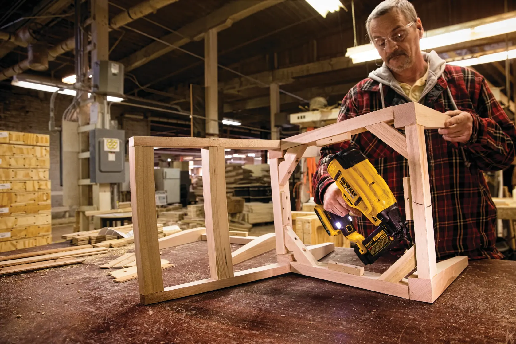 Una persona utiliza una clavadora DEWALT DCN680B para ensamblar un marco de madera en un taller. El rostro de la persona está desenfocado y lleva una camisa de cuadros. El espacio de trabajo está lleno de madera y equipos de taller.