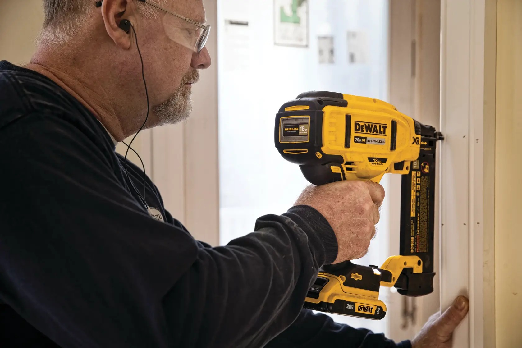 Una persona utiliza una clavadora inalámbrica DEWALT DCN680B para fijar una moldura a un marco de puerta. La herramienta de marca amarilla y negra se sostiene con una mano, y la cara de la persona está difuminada para la privacidad.