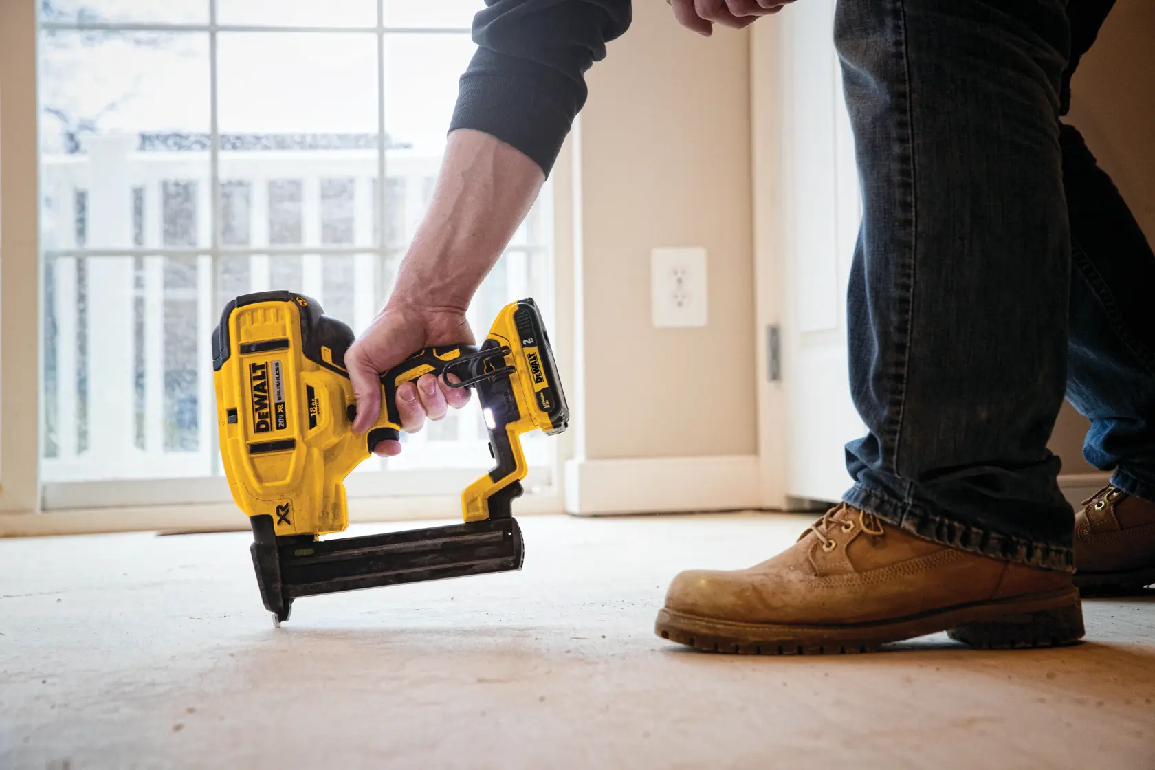 Una persona sostiene y utiliza una clavadora inalámbrica DEWALT sobre un suelo, con una ventana y una puerta al fondo.