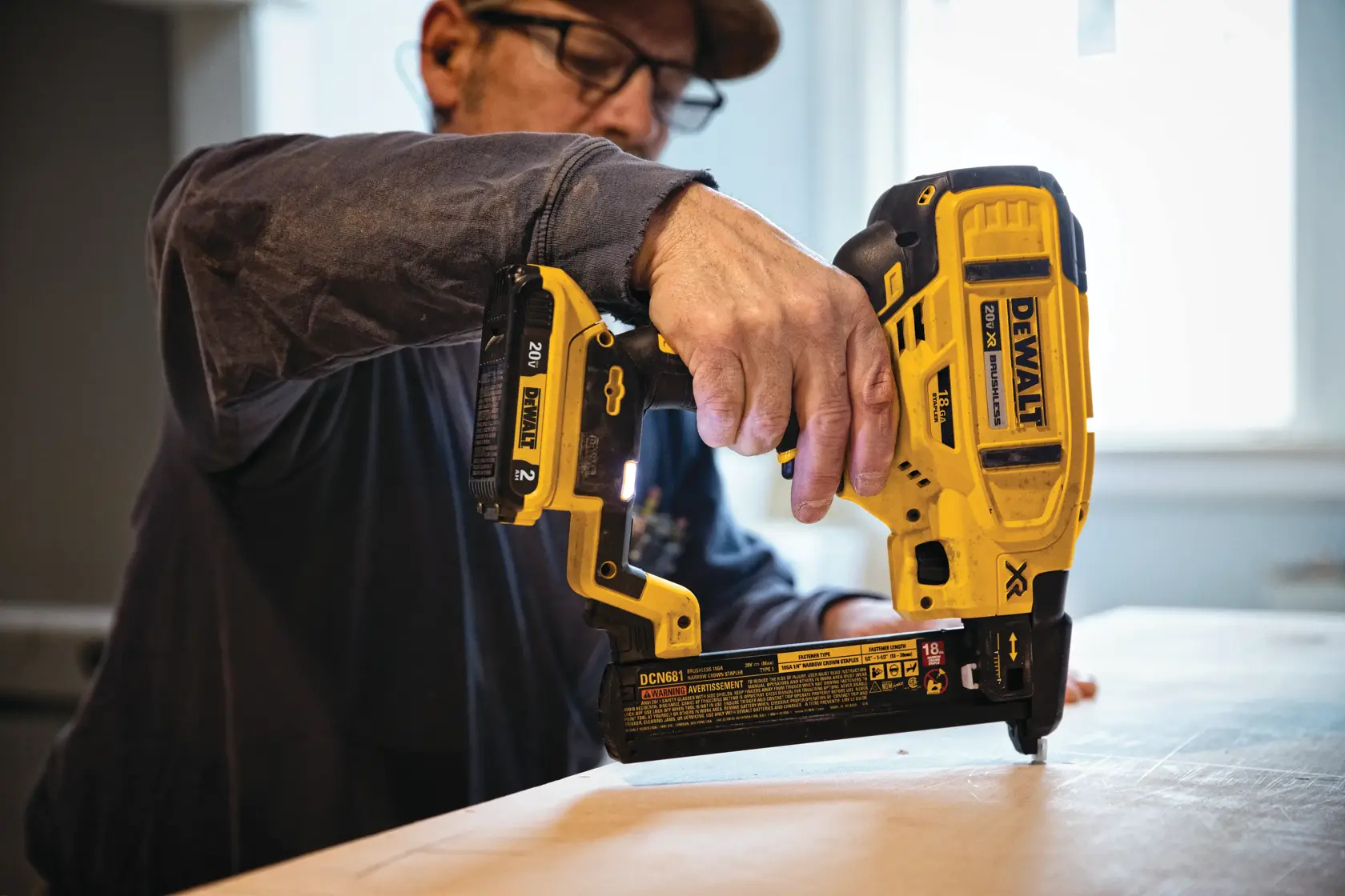 Una persona utiliza una clavadora inalámbrica amarilla DEWALT DCN681B para fijar madera en una superficie de trabajo, en interiores.