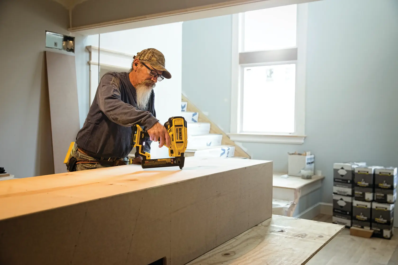 Una persona está usando una clavadora DEWALT DCN681B para ensamblar madera en un espacio de trabajo interior. Se ven cajas de materiales en el fondo y la persona lleva ropa de trabajo y una gorra.
