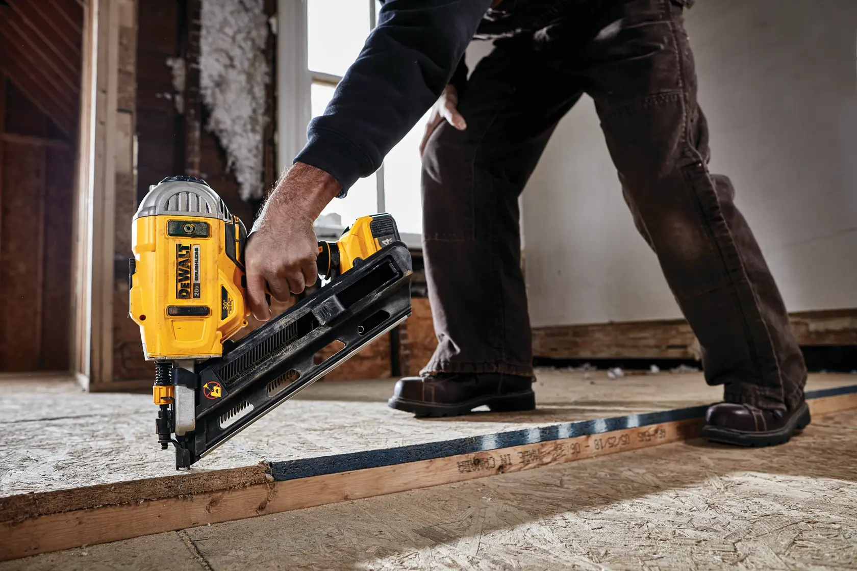 Una persona utiliza una clavadora inalámbrica DEWALT DCN692B para fijar una viga de madera durante trabajos de construcción en interiores. La herramienta es amarilla y negra, y la persona lleva pantalones oscuros y zapatos marrones.