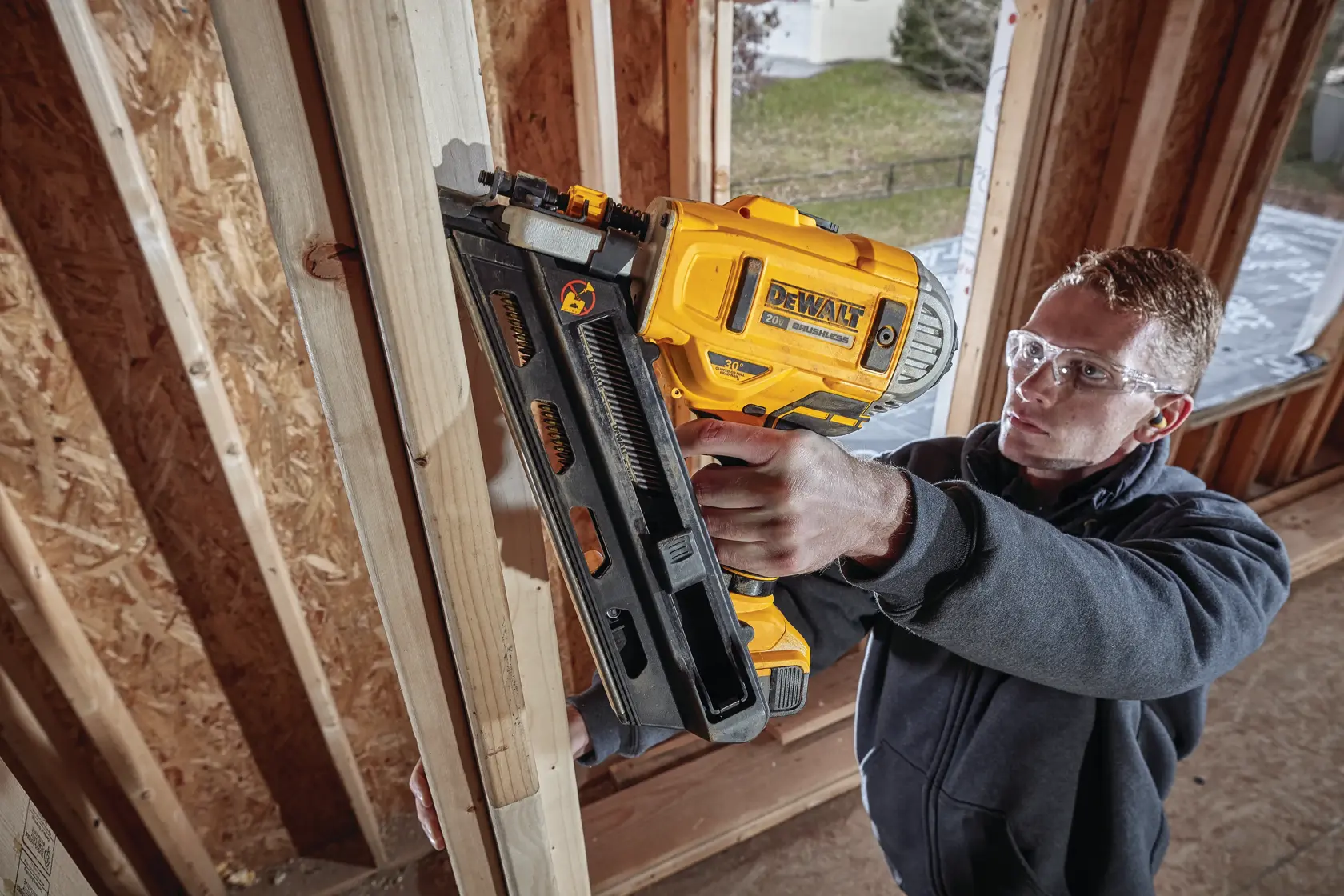 Una persona está usando una clavadora inalámbrica DEWALT DCN692B para fijar montantes de madera dentro de un edificio en construcción. El rostro de la persona está difuminado por privacidad. La escena es en el interior y muestra una estructura de madera sin terminar.