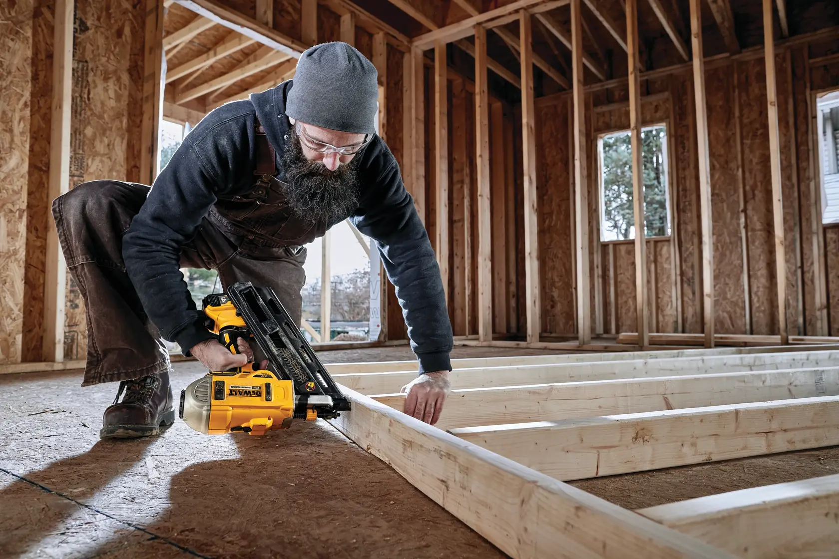 Una persona utiliza una pistola de clavos amarilla DEWALT DCN692B para fijar una estructura de madera dentro de una casa en construcción.