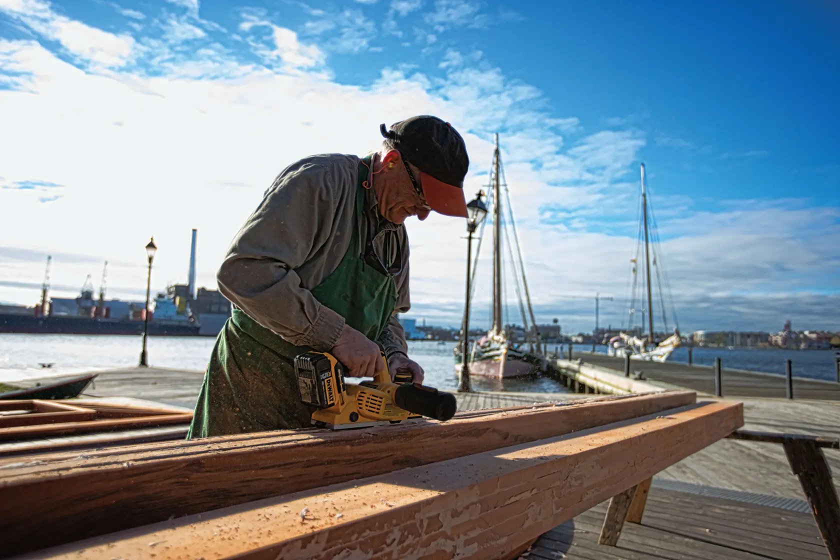 Una persona está usando una herramienta eléctrica DEWALT DCP580B para cepillar una viga de madera al aire libre cerca de un muelle, con barcos y edificios industriales al fondo. El rostro de la persona está difuminado.