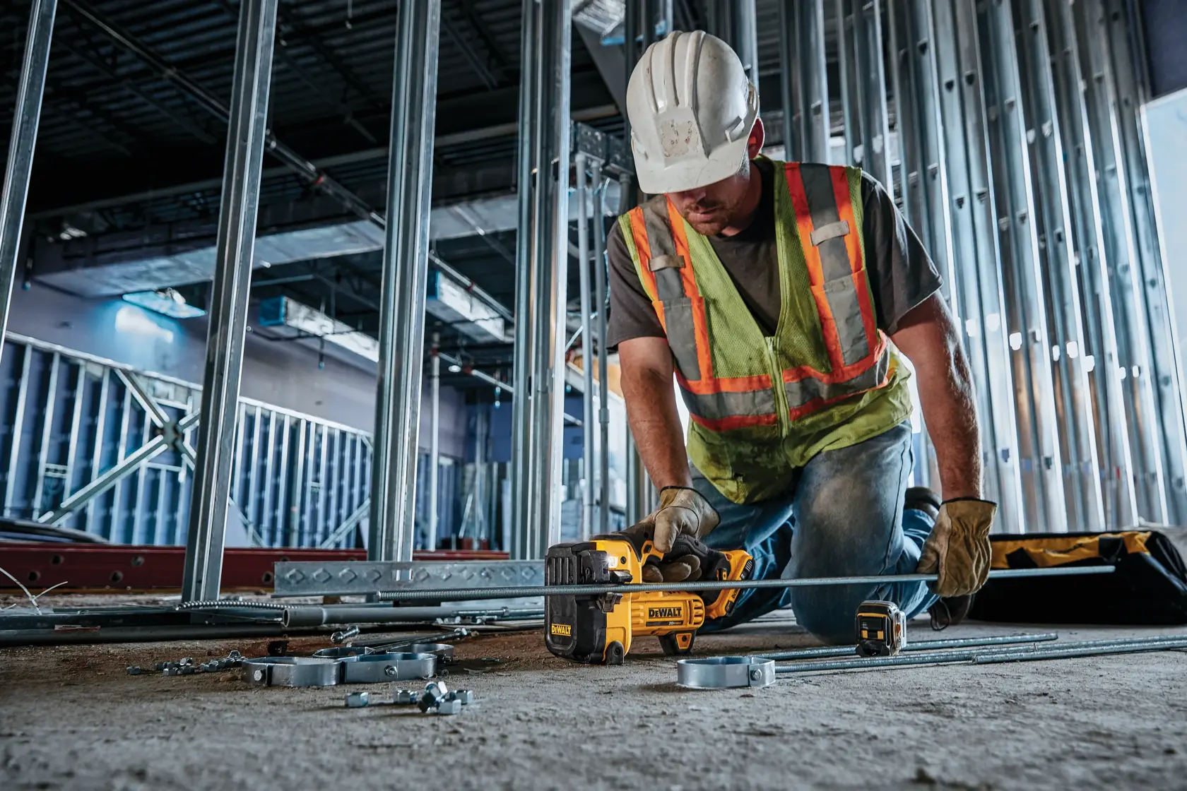 Un trabajador de construcción con chaleco de seguridad, guantes y casco utiliza una herramienta DEWALT DCS350B para cortar barras de metal en un sitio de construcción, rodeado de estructuras metálicas y materiales de obra.