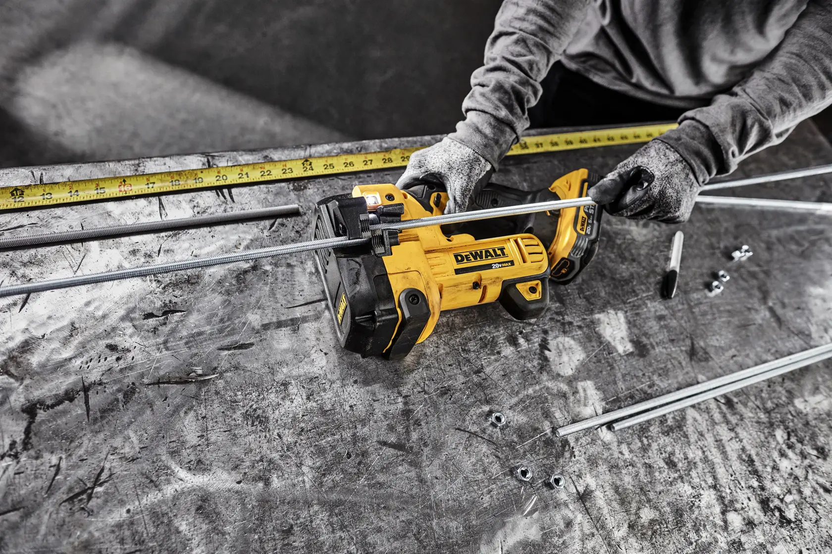 Una persona con guantes utiliza un cortador de varillas roscadas inalámbrico amarillo DEWALT para cortar varillas metálicas sobre una mesa de trabajo. Se observan varias varillas, tuercas y una cinta métrica en la mesa.