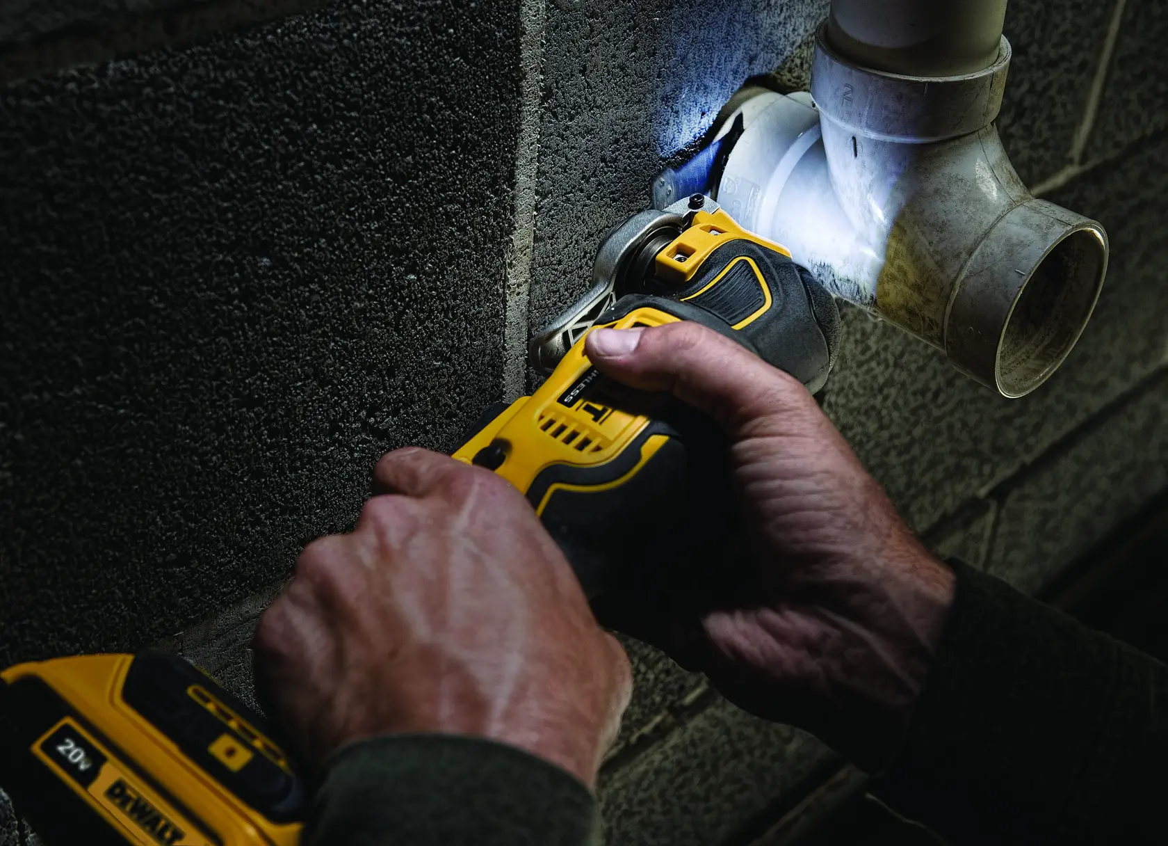 Primer plano de manos utilizando una herramienta multiusos oscilante inalámbrica DEWALT para cortar una pared de concreto cerca de una tubería de plástico.