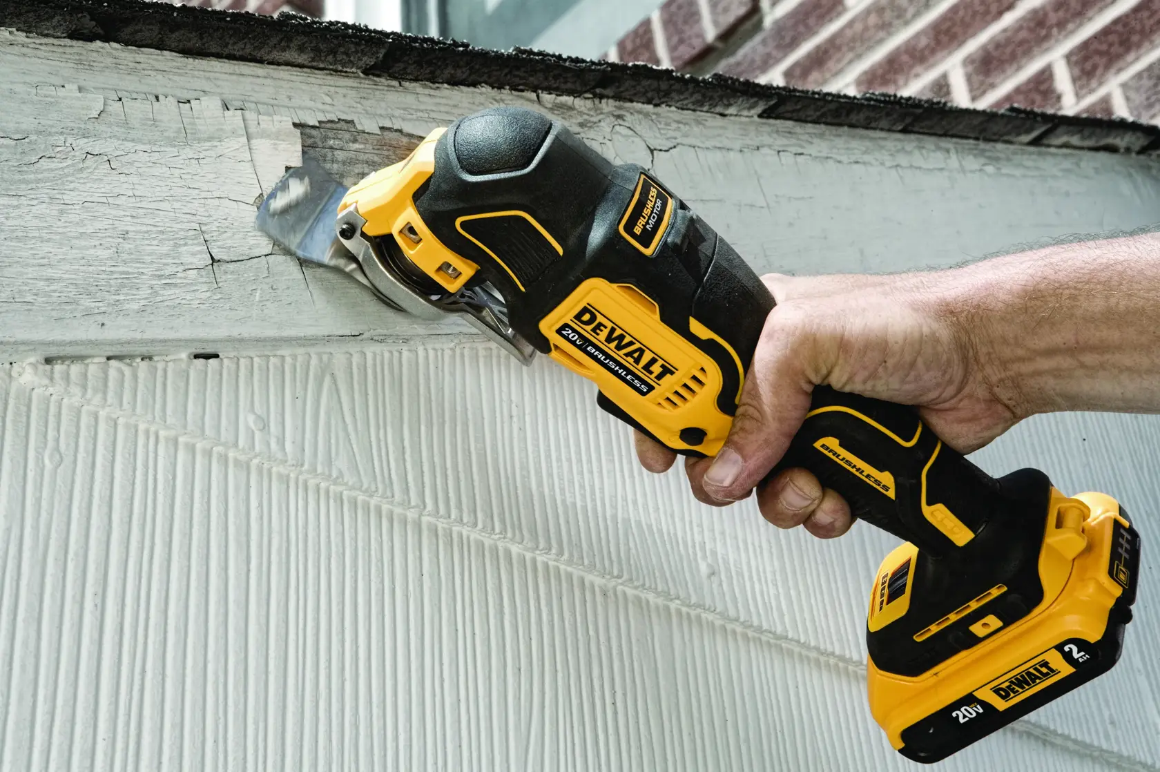 Una mano sostiene una herramienta multifunción oscilante inalámbrica DEWALT DCS356B que se está usando para raspar pintura vieja de una superficie de madera.