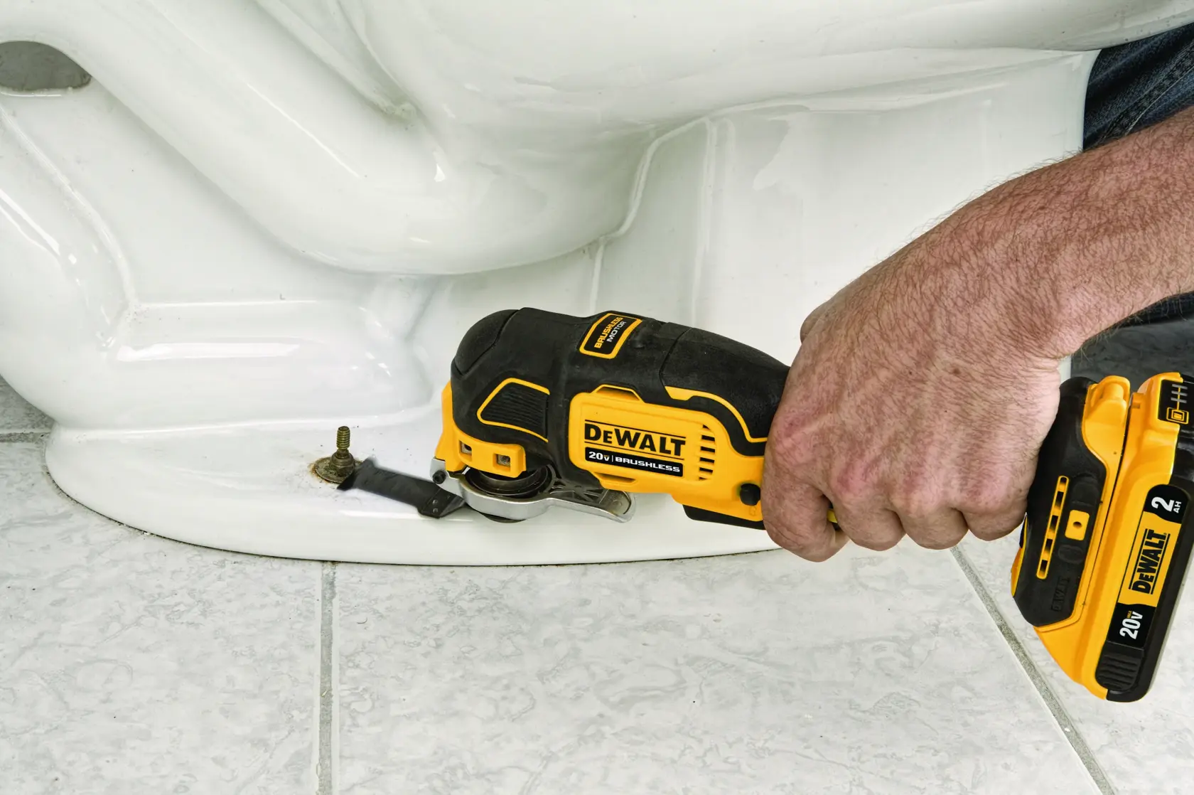 Una persona utiliza una herramienta multifuncional oscilante inalámbrica DEWALT DCS356B 20V para cortar o remover un perno cerca de la base de un inodoro de cerámica blanca sobre un suelo de baldosas.