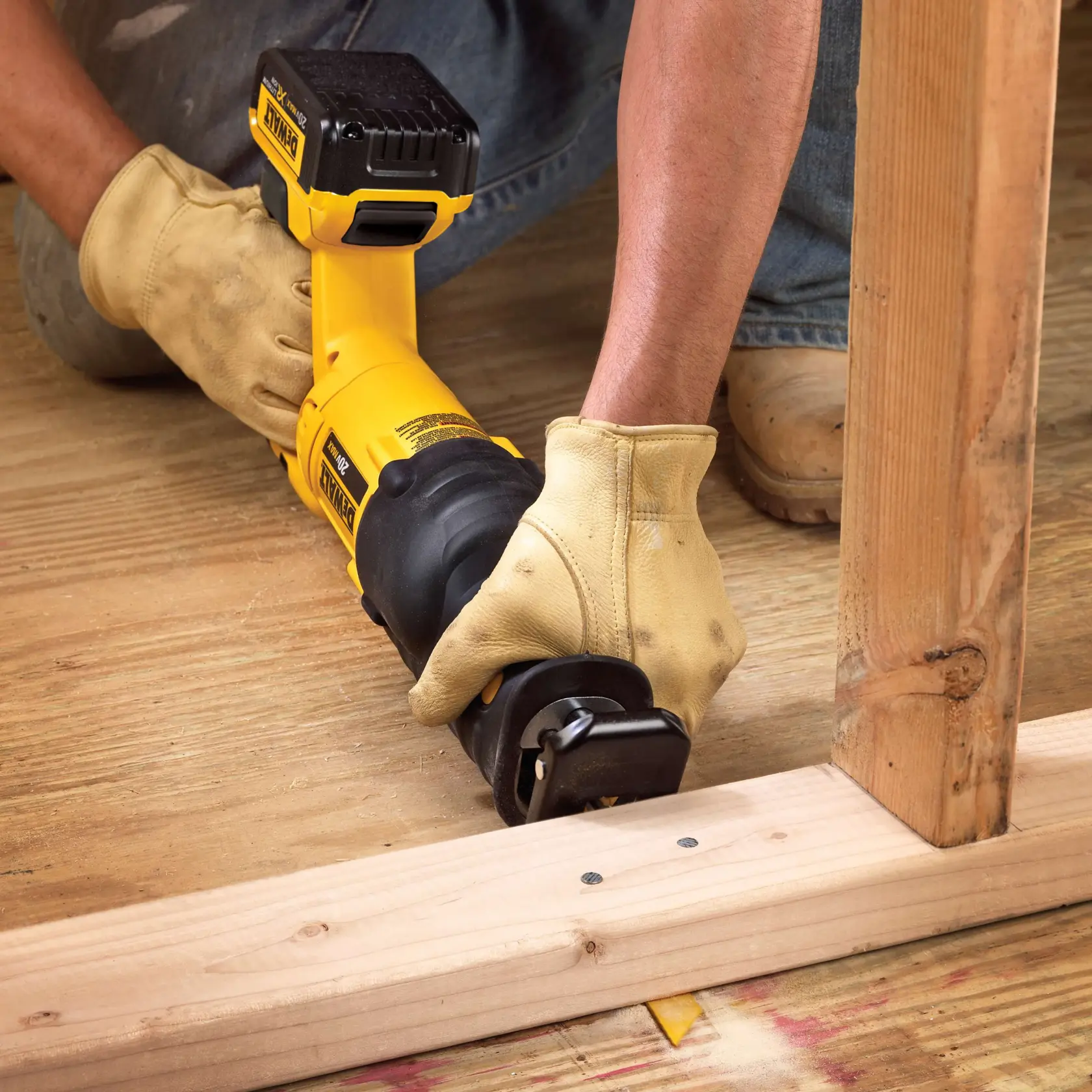Una persona con guantes de trabajo amarillos está usando una sierra recíproca inalámbrica DEWALT amarilla para cortar madera en un sitio de construcción. La sierra está posicionada cerca de una viga de soporte de madera y un suelo de tablones de madera.