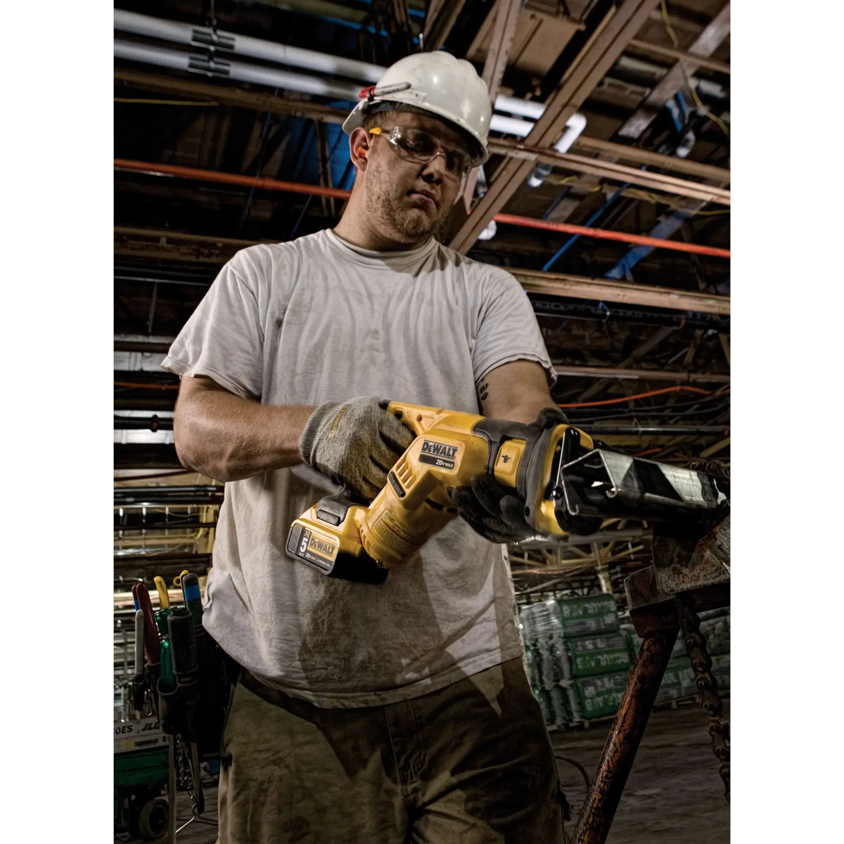Una persona con guantes de trabajo y casco protector está usando una sierra recíproca DEWALT DCS387B en un entorno industrial. Al fondo se ven materiales de construcción y tuberías de techo expuestas.