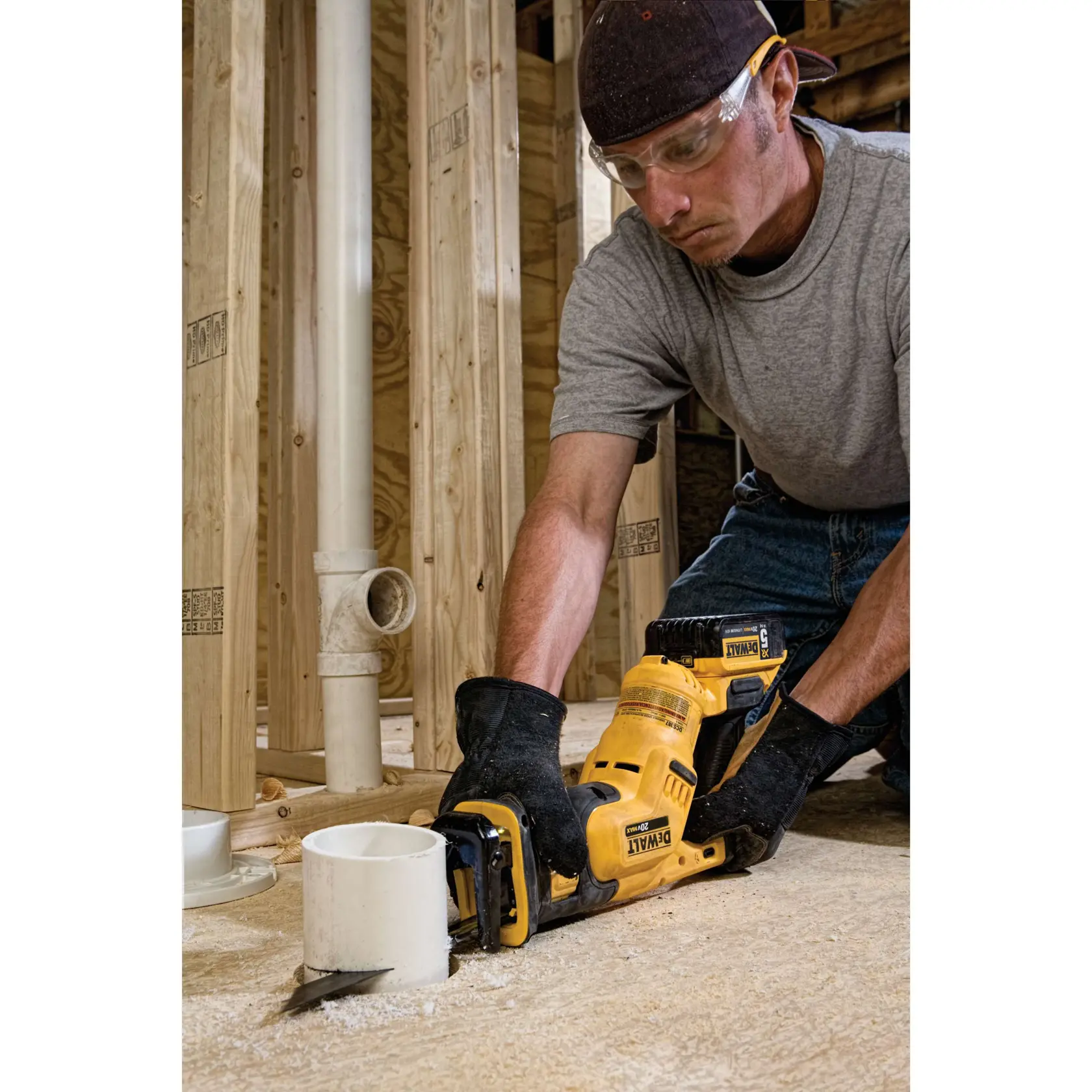 Una persona con guantes y gorra está utilizando una sierra recíproca inalámbrica DEWALT para cortar un tubo de PVC en una obra de construcción con armazón de madera y tuberías visibles.