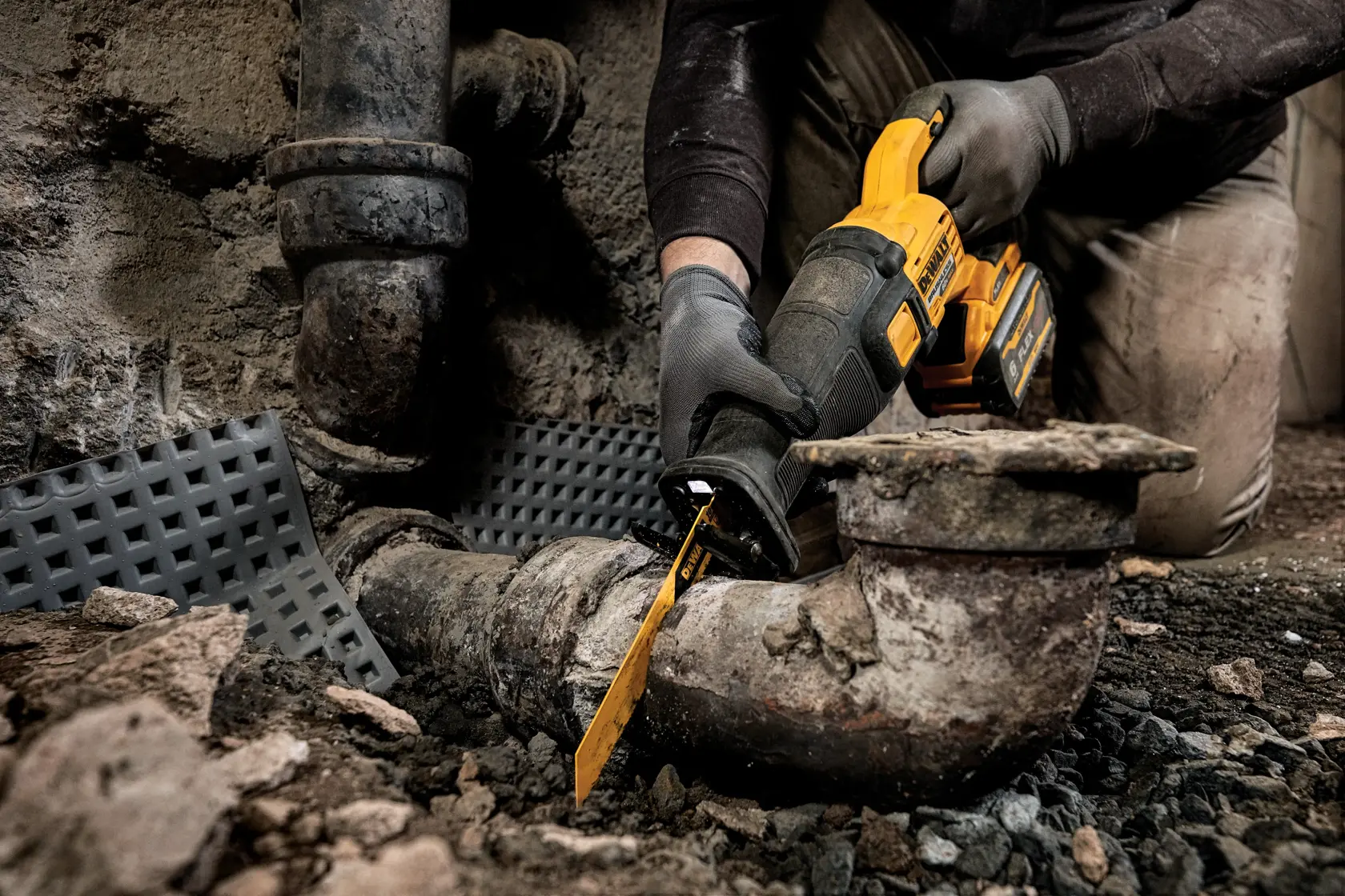 Una persona con guantes utiliza una sierra recíproca inalámbrica DEWALT para cortar una tubería de metal vieja en un entorno industrial y áspero. El área está polvorienta con escombros en el suelo.