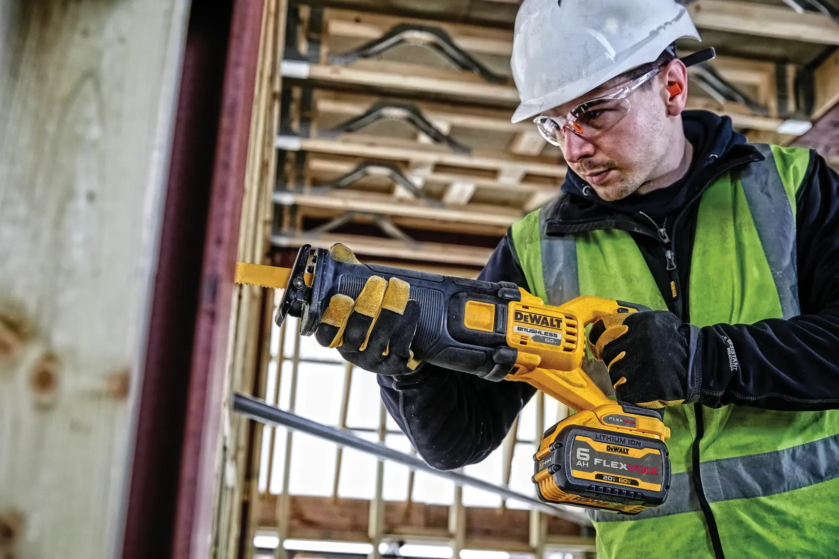 Una persona vestida con chaleco de seguridad, guantes y casco usa una sierra recíproca inalámbrica DEWALT (DCS389B) en un entorno de construcción. La sierra cuenta con una batería FLEXVOLT y está situada cerca de vigas de madera.