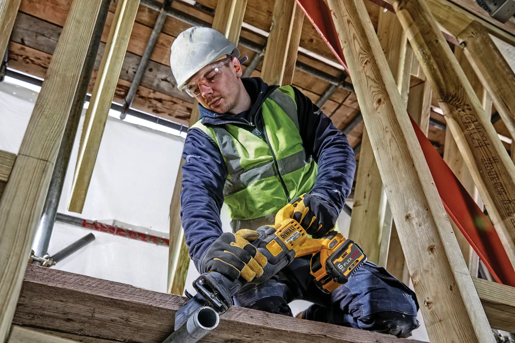 Un trabajador de la construcción que lleva chaleco de seguridad, guantes y casco usa una sierra recíproca inalámbrica DEWALT (modelo DCS389B) para cortar una tubería de metal en una obra de madera.