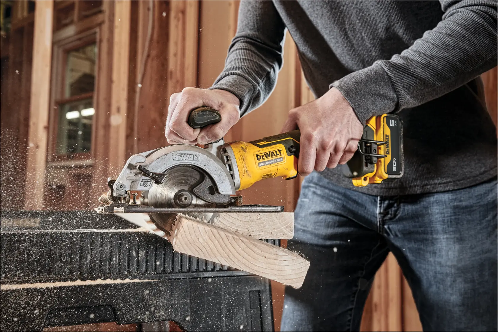 Una persona usa una sierra circular inalámbrica DEWALT DCS571B para cortar un trozo de madera en un taller. Se puede ver el aserrín volando mientras la hoja corta la madera.