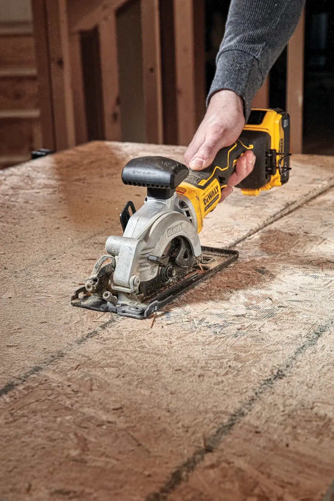 Una persona utiliza una sierra circular inalámbrica DEWALT para cortar una hoja de madera sobre una mesa de trabajo en un taller.