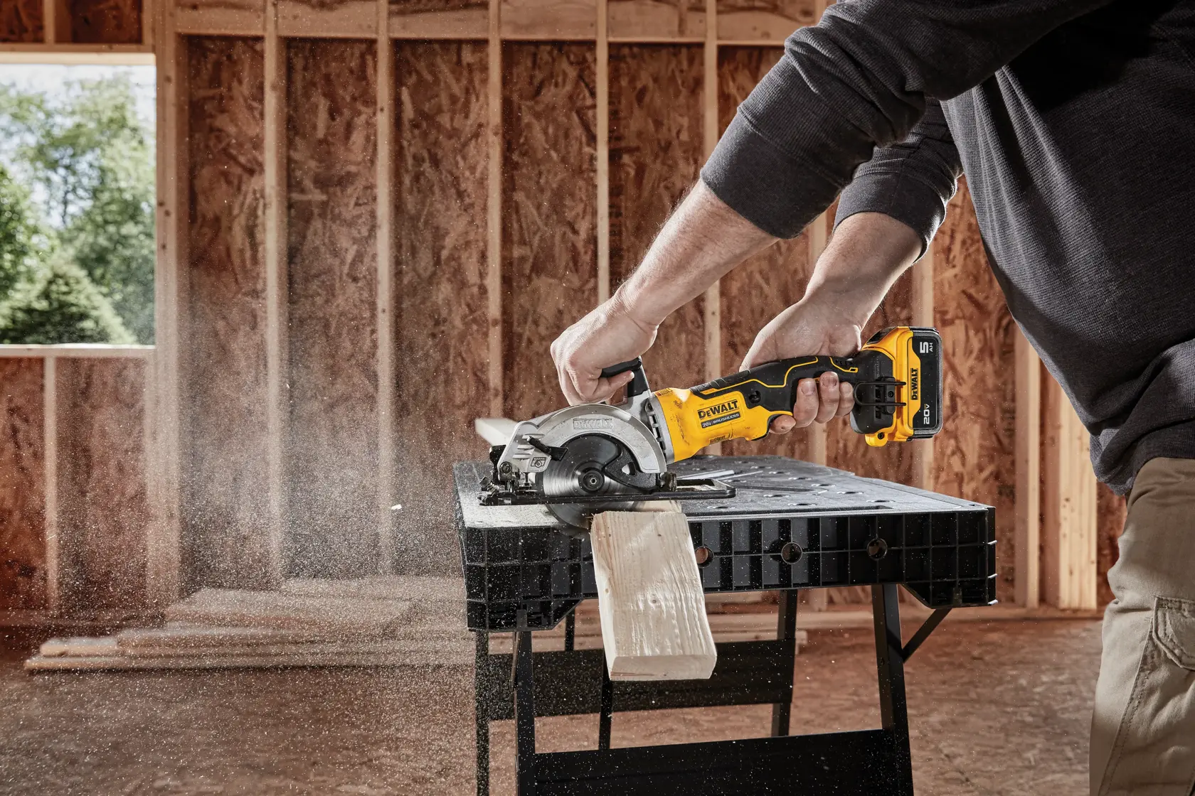 Una persona usa una sierra circular inalámbrica DEWALT para cortar un trozo de madera sobre un banco de trabajo dentro de un edificio en construcción.